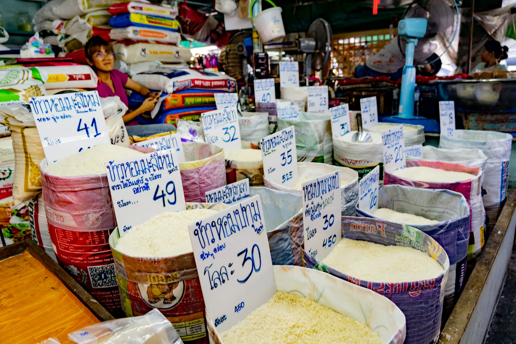 写真はタイのバンコクの市場で撮影されたもので、様々な種類の米が売られている様子を捉えています。手前には、米が袋や容器に山盛りに入れられ、それぞれに手書きのタイ語の価格表示札が立てられています。札には「ข้าวหอมมะลิใหม่」（カオホムマリマイ、ジャスミンライス）などの米の種類や、キロあたりの価格が記載されています。価格は30～49バーツ程度です。  奥には、商品を売る女性が座っており、スマートフォンを操作しているように見えます。背景には米袋が積み上げられ、扇風機やその他の商品が並んでいます。全体的に活気のある市場の雰囲気が出ており、生活感あふれる風景です。