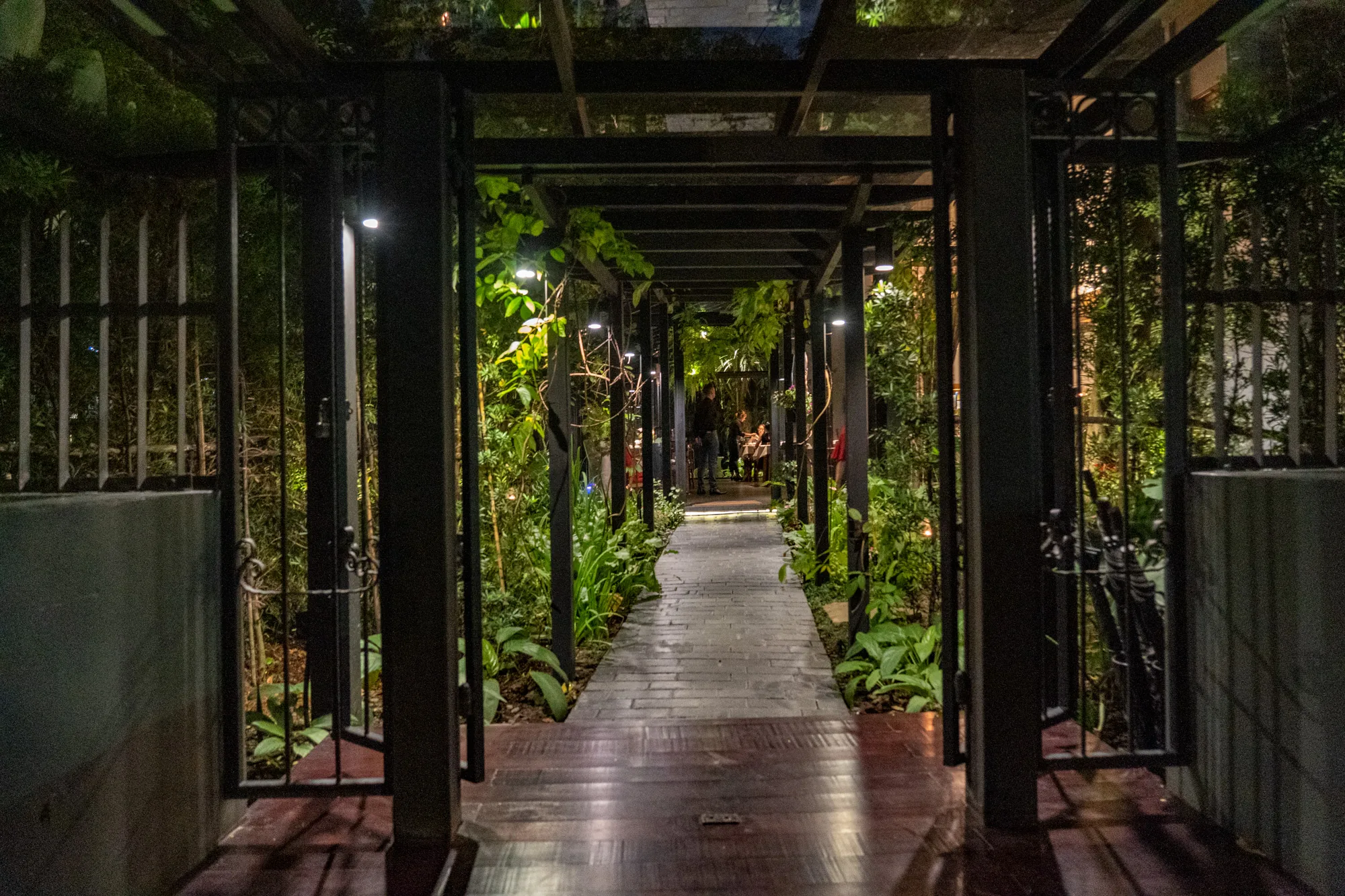写真は、タイのバンコクで撮影された、夜の庭園の通路を写したものです。視点は、通路の入り口にあり、両側に開いた鉄製の装飾的な門があります。通路はレンガで舗装されており、暗い木製の床から始まっています。  通路の上には、金属製の格子状の屋根があり、それが植物によって覆われています。屋根には等間隔に小さなライトが設置されており、通路を照らしています。通路の両側には、さまざまな種類の緑豊かな植物が生い茂っており、熱帯の雰囲気を醸し出しています。  通路の奥には、テーブルを囲んで談笑している人々の姿が見えます。そのため、ここはレストランかバーの一部である可能性があります。全体的に、写真は静かでリラックスした雰囲気で、夜のバンコクの庭園の美しさを捉えています。