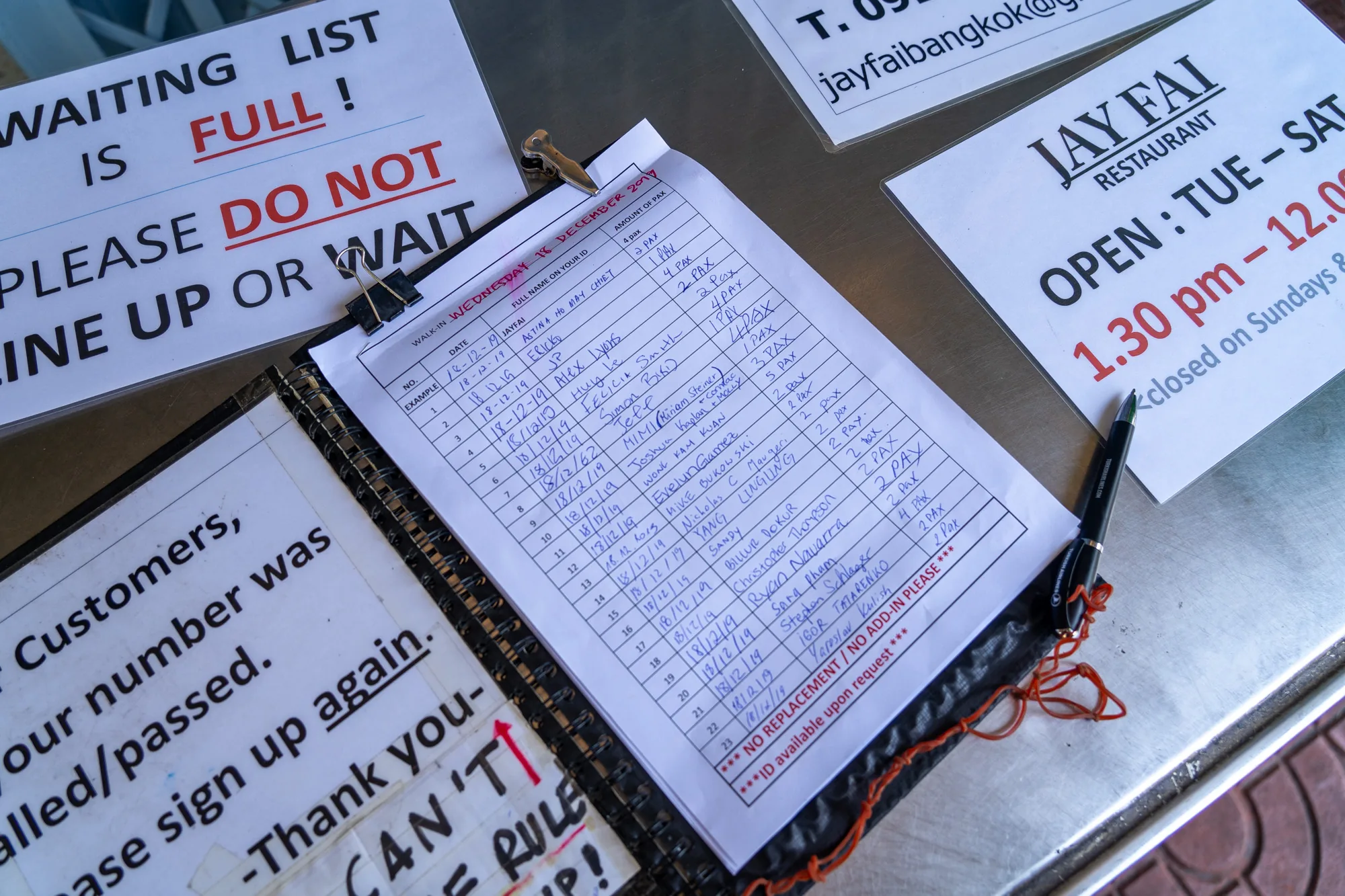 この写真は、タイのバンコクにある有名な屋台料理店「ジェイ・ファイ (Jay Fai)」のウェイティングリストを写したものです。写真には、順番待ちの顧客向けに書かれた複数の案内が写っています。  まず、大きな文字で「WAITING LIST IS FULL! PLEASE DO NOT LINE UP OR WAIT」と書かれた紙が目に入ります。これは、ウェイティングリストがいっぱいで、並んだり待ったりしないように顧客に伝えるものです。  次に、テーブルの上に置かれたバインダーのウェイティングリストが写っています。リストは手書きで、日付、顧客の名前、人数が記載されています。リストの日付は「WEDNESDAY 18 DECEMBER 2019」となっています。また、リストの下部には「*** NO REPLACEMENT / NO ADD-IN PLEASE *** ***ID available upon request ***」と書かれています。  さらに、レストランの営業時間を示す看板も写っています。「JAY FAI RESTAURANT OPEN : TUE – SAT 1.30 pm - 12.00 closed on Sundays &」と記載されており、火曜日から土曜日の午後1時30分から午前0時まで営業していることが分かります。  写真全体から、非常に人気のあるレストランであることが伺えます。ウェイティングリストが常にいっぱいで、多くの観光客が訪れている様子が想像できます。