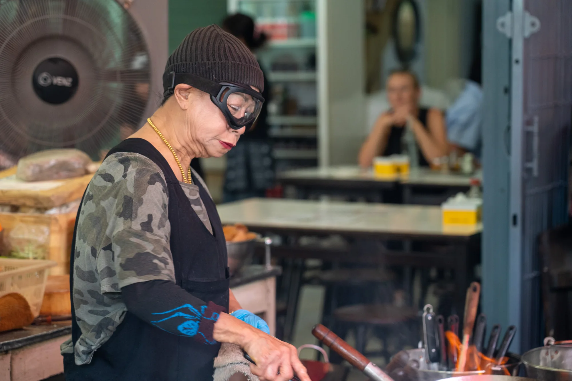 写真はタイのバンコクで撮影された、屋台料理店の様子を捉えたものです。手前にいるのは、調理をしている年配の女性です。彼女は黒いニット帽をかぶり、ゴーグルをしています。迷彩柄のTシャツに黒いエプロンをつけ、首には金のネックレスをしています。手には青い手袋を着用しており、鍋を操る手元が写っています。鍋は火にかけられ、炎が見えます。背景には、テーブルと椅子があり、一人の女性客が座っています。店内の様子が伺え、ローカルな雰囲気が伝わってきます。また、扇風機や食材などが置かれており、屋台の活気を感じさせる一枚です。