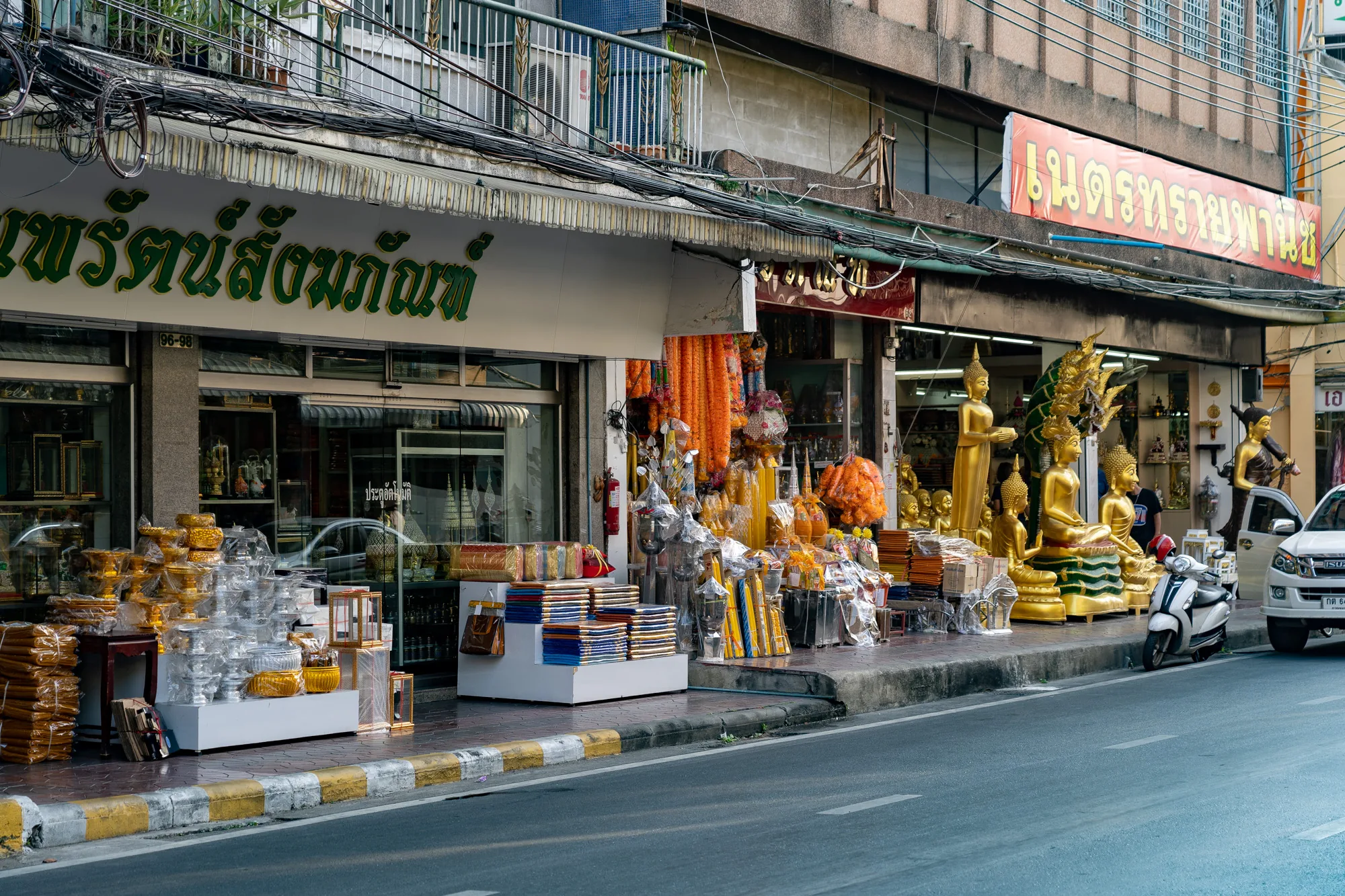写真はタイのバンコクで撮影された街の風景です。画面手前には道路があり、その奥には歩道が続いています。歩道沿いには複数のお店が並び、それぞれのお店が仏具や仏像などを販売しています。  左端のお店では、金色の器や銀色の器などが積み上げられており、その多くが透明なビニールで包装されています。隣の店では、オレンジ色の花輪や、カラフルな布が積まれた棚などが見えます。右側の店では、さまざまなポーズをとった金色の仏像が並んでおり、それぞれ独特の装飾が施されています。  歩道には、白いスクーターと白いピックアップトラックが駐車されています。背景には、複雑に絡み合った電線が張り巡らされており、建物の壁にはタイ語の看板が掲げられています。全体的に落ち着いた雰囲気で、バンコクの日常風景を捉えた写真です。