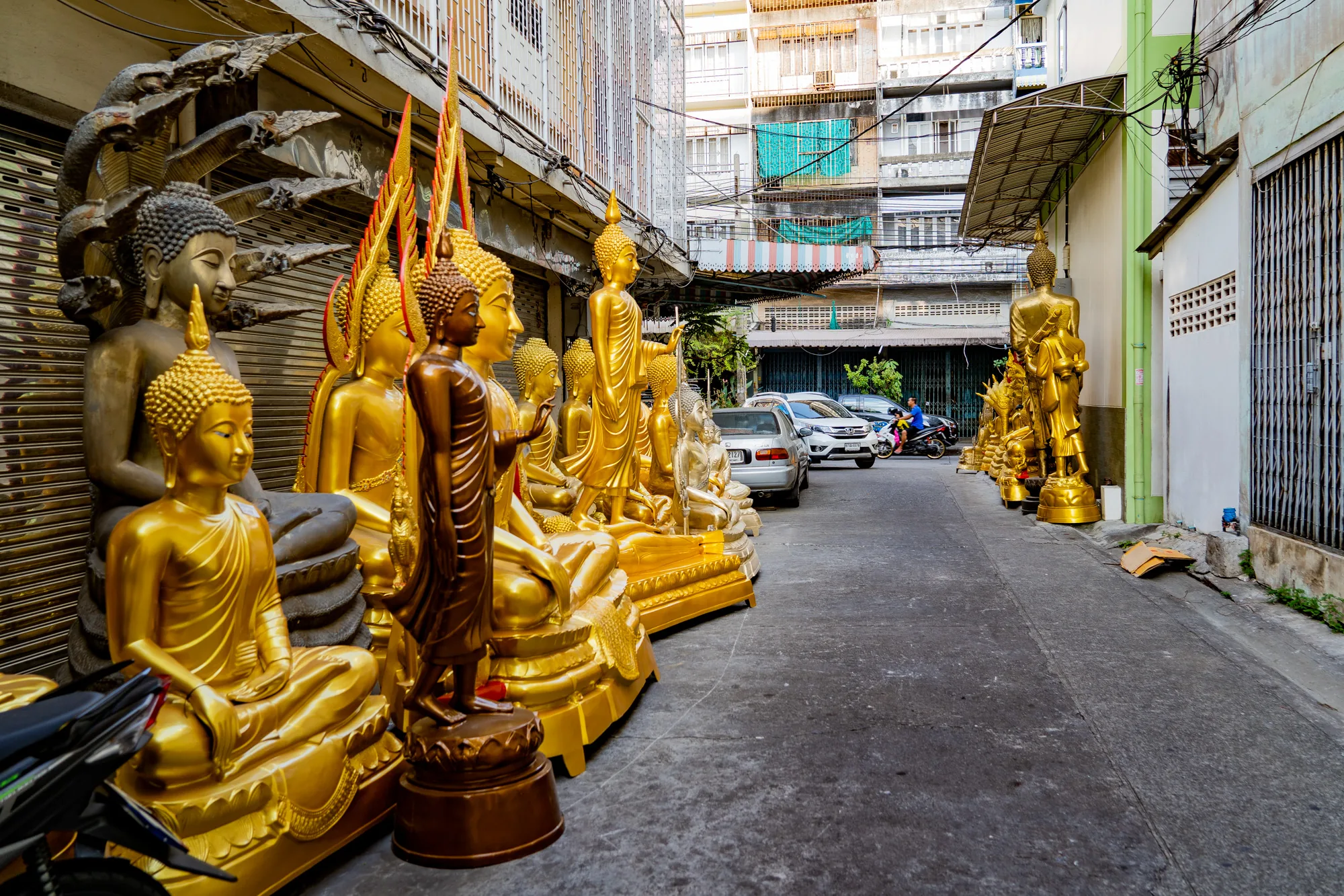 写真はタイのバンコクで撮影された、路地の風景を捉えています。路地の片側には、様々な大きさや種類の仏像がずらりと並んでいます。仏像は金色に輝いており、座禅を組んだ姿や、立ち姿など、多様なポーズをしています。背景には、古びた建物が立ち並び、電線が複雑に絡み合っています。路地には車やバイクが停まっており、バイクに乗った人が通り過ぎる様子も見えます。路地は少し薄暗く、生活感が漂っています。全体的に、タイの日常風景と仏教文化が混ざり合った独特の雰囲気が感じられる写真です。