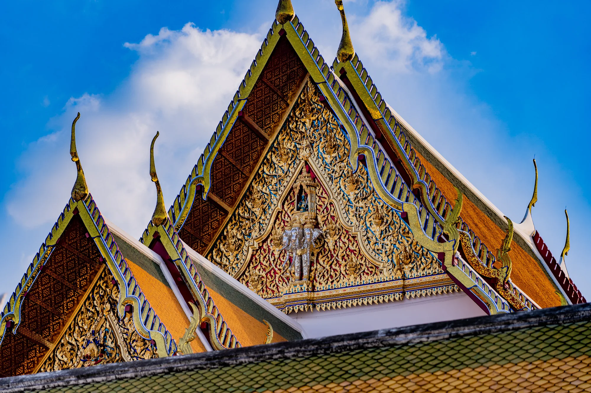 タイのバンコクで撮影された写真です。青空を背景に、タイの寺院の屋根が写っています。屋根は伝統的なタイ様式で、金色を基調とした装飾が細部にまで施されています。屋根の先端は、上向きに反り返った形状をしており、金色に輝いています。屋根の表面には、複雑な模様が彫り込まれており、豪華絢爛な雰囲気を醸し出しています。空には白い雲が浮かんでおり、青空とのコントラストが美しいです。写真全体から、タイの寺院の壮麗さと繊細さを感じることができます。