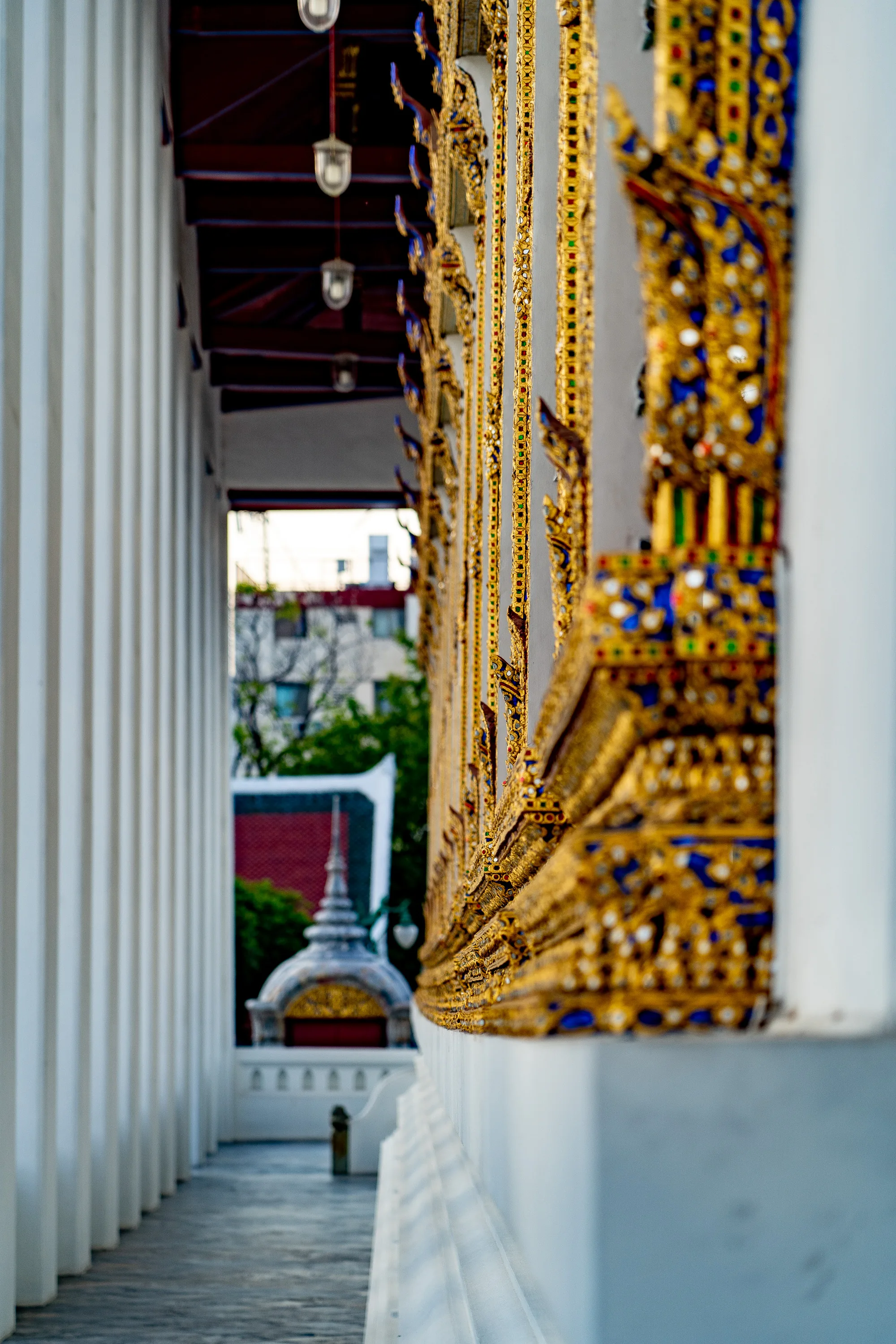 この写真はタイのバンコクで撮影されたもので、寺院の装飾が特徴的です。画面右側には、白壁に施された金色と青色の装飾が精巧に配置されています。これらの装飾は、伝統的なタイの様式を反映しており、細部にわたる美しい模様が目を引きます。左側には、白い柱が整然と並び、奥行きを強調しています。柱の奥には、別の建物の屋根の一部と、その背景に緑の木々と建物が見え、市街地にある寺院であることを示唆しています。全体的に、厳かで華やかな雰囲気が漂い、タイの文化的な魅力を伝えています。