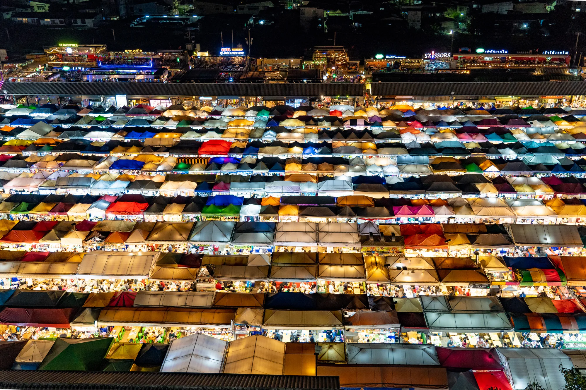 写真はタイのバンコクで撮影された夜の市場の鳥瞰図です。数えきれないほどのテントが密集して並び、カラフルな屋根が光を反射して、まるで宝石箱のように輝いています。赤、青、緑、黄色など様々な色のテントがランダムに配置され、活気あふれる雰囲気を醸し出しています。テントの下には人々が集まり、食事をしたり、買い物をしたりしている様子が伺えます。  写真の上部には、市場を取り囲む建物やお店の看板が見えます。これらの建物は市場よりも少し高く、夜空にシルエットとして浮かび上がっています。全体的に、写真はバンコクの夜の活気とエネルギーを見事に捉えており、観光客や地元の人々が集まる賑やかな場所であることが伝わってきます。