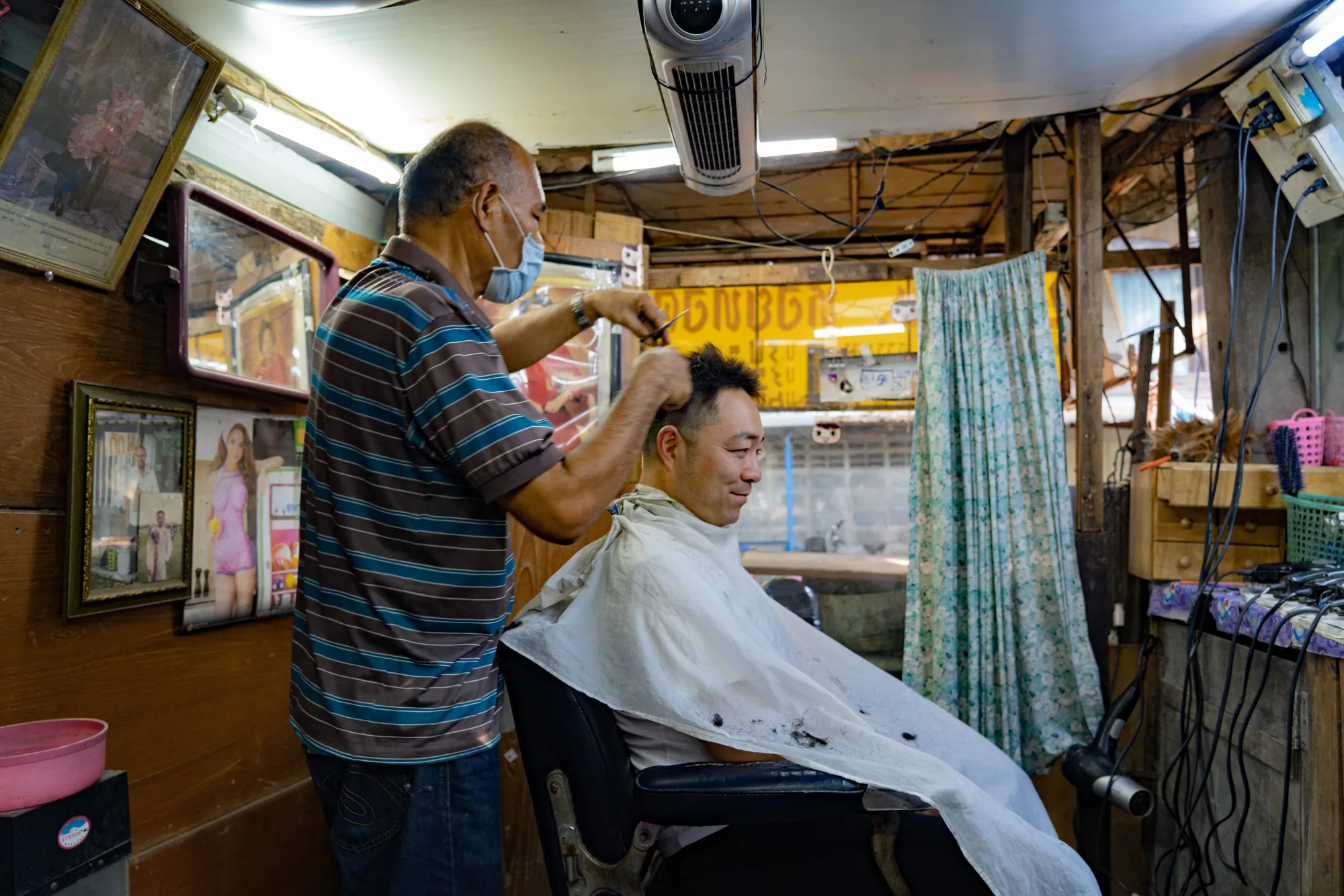 写真はタイのバンコクで撮影された、ローカルな雰囲気の床屋さんの様子を捉えています。店内の様子は少し雑然としており、壁には複数の写真や鏡が飾られています。理容師と思われる男性が、マスクを着用して顧客の髪を切っています。顧客は理容椅子に座り、ケープをかけています。全体的に、生活感あふれる、親しみやすい雰囲気の写真です。天井からは扇風機が吊り下げられ、壁には電線やコンセントがむき出しになっています。