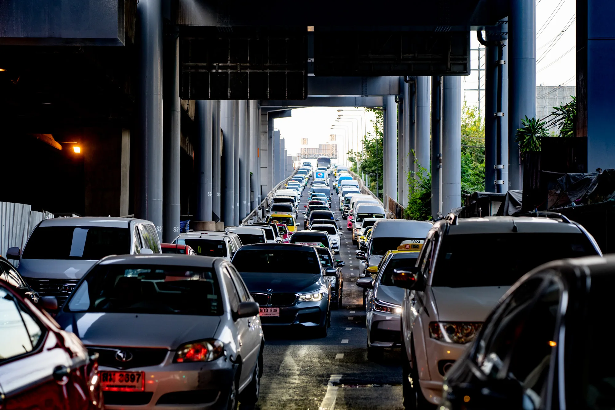 写真はタイのバンコクで撮影されたもので、高架道路の下で発生している激しい交通渋滞の様子を捉えています。画面奥に向かって、車の列が何台も連なっており、渋滞の深刻さを物語っています。手前の車両は画面を遮るように配置され、迫りくるような圧迫感を与えています。車種は様々で、タクシーと思われる黄色い車体も見られます。背景には、コンクリート製の高架道路の支柱が整然と並び、都市のインフラを感じさせます。薄暗い高架下と、遠くに見える明るい空とのコントラストが、写真に奥行きを与えています。全体的に、バンコクの都市生活における交通問題の深刻さを物語る一枚と言えるでしょう。