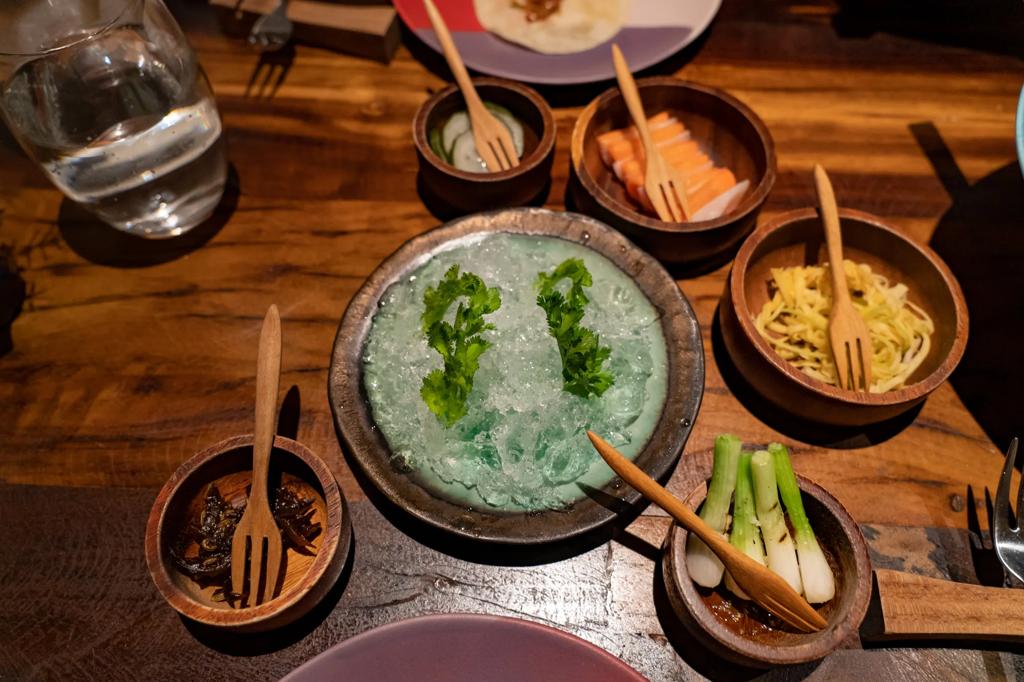 写真は、タイのバンコクで撮影されたと思われる、コース料理の一場面を捉えています。木製のテーブルの上に、様々な料理が盛り付けられた小さめの器が並んでいます。中央の皿には、砕いた氷の上にパクチーが飾られており、涼しげな印象を与えます。周りには、キュウリの酢漬け、サーモン、麺料理、ネギ焼き、海藻のような料理がそれぞれ木製の小鉢に盛り付けられ、木製のミニフォークが添えられています。これらの料理は、コース料理の一品ずつであると考えられます。上品な雰囲気で、特別な食事の体験を想起させます。テーブルの材質や食器の質感から、高級感も感じられます。