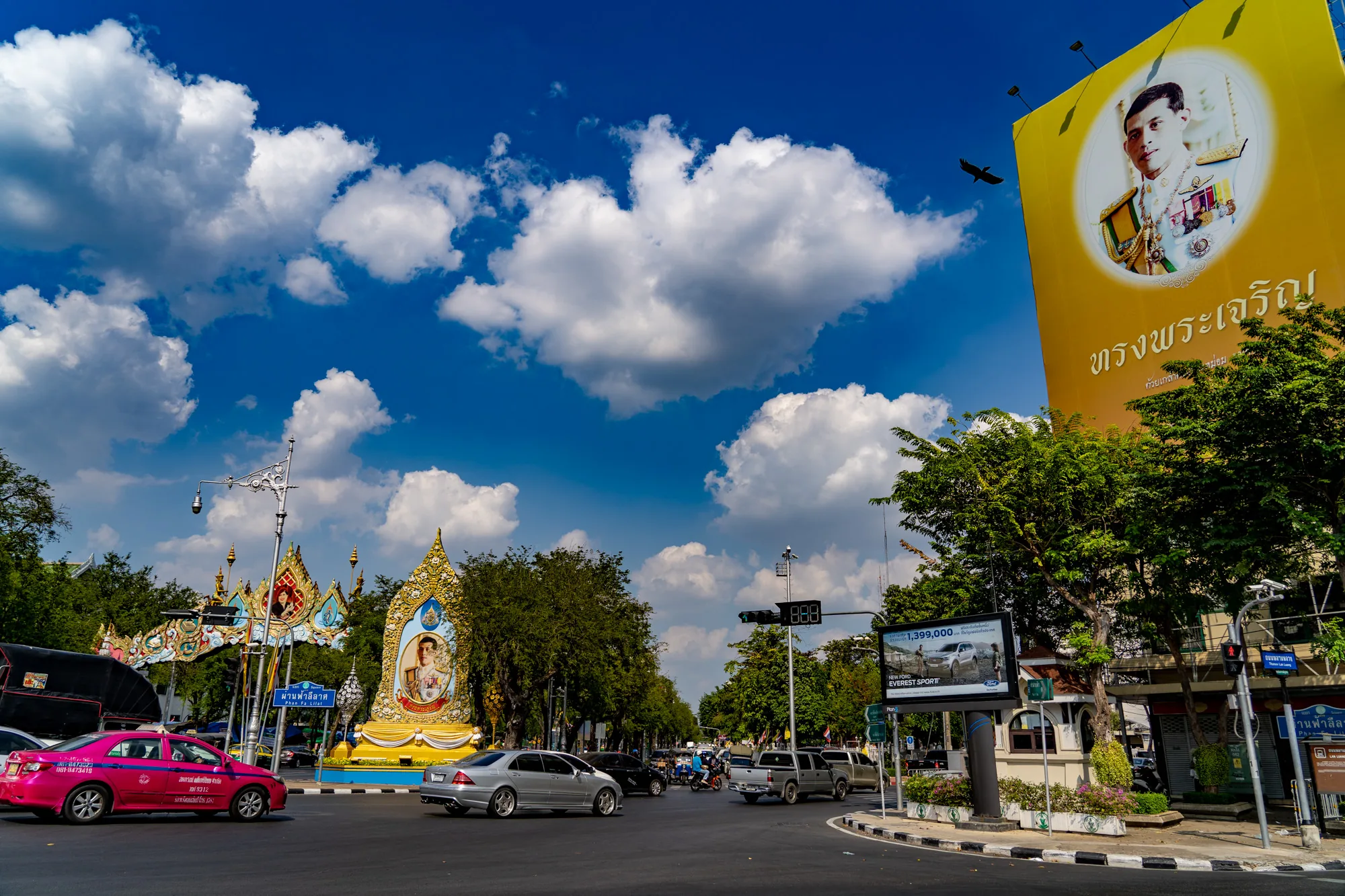 写真はタイのバンコクで撮影されたもので、都市の賑やかな様子が写されています。青空には白い雲が広がり、その下には交通量の多い道路が見えます。道路にはピンク色のタクシーやシルバーの乗用車などが走っており、交通標識や信号機も設置されています。道路沿いには街路樹が並び、緑豊かな風景を作り出しています。  特に目を引くのは、道路の左右に設置された巨大な王様の肖像画です。黄色い背景に王様の写真が大きく描かれており、タイの文化や信仰を象徴しているようです。また、道路標識や広告看板なども写っており、都市の喧騒を伝えています。全体的に明るく鮮やかな色彩で、活気あふれるバンコクの雰囲気が伝わってくる写真です。
