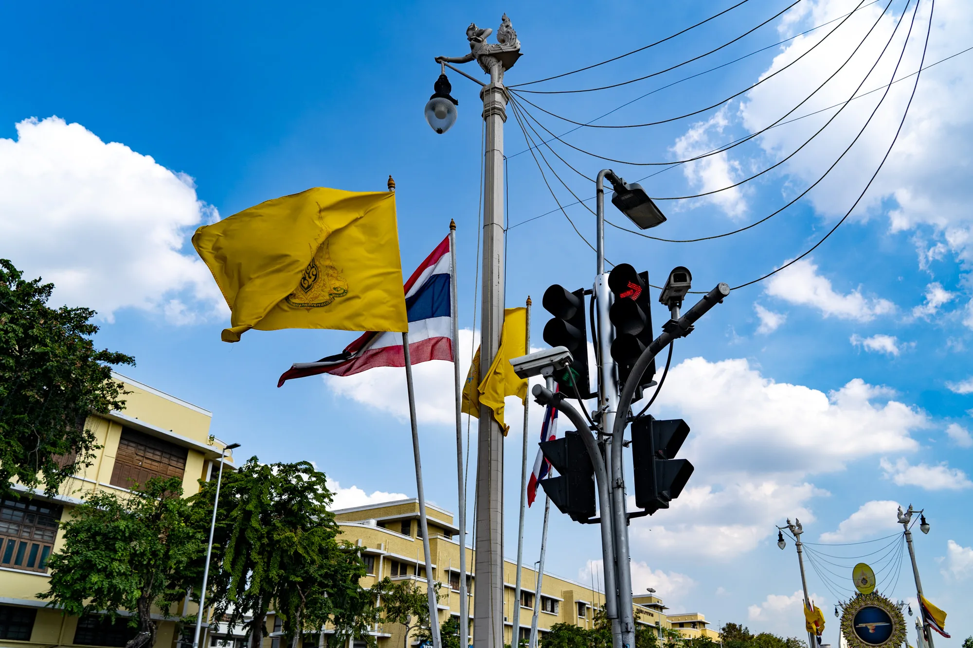 写真はタイのバンコクで撮影されたもので、鮮やかな青空と白い雲が広がる背景に、複数の旗がはためいている様子が写っています。一番手前には大きな黄色の旗があり、旗にはタイ王室の紋章が描かれています。その奥にはタイの国旗であるトリコロール（赤・白・青・白・赤）が風になびいています。他にも黄色い旗が見えます。  画面右側には交通信号機があり、赤い矢印が表示されています。信号機の支柱には監視カメラが取り付けられています。電線が空を横切り、複雑な模様を描いています。  背景には、淡い黄色の建物が写っています。建物は複数階建てで、伝統的なタイの建築様式を取り入れています。手前には緑の木々が茂っており、写真全体に自然の要素を加えています。全体的に、タイの活気ある都市の風景と文化的な要素が組み合わさった写真です。