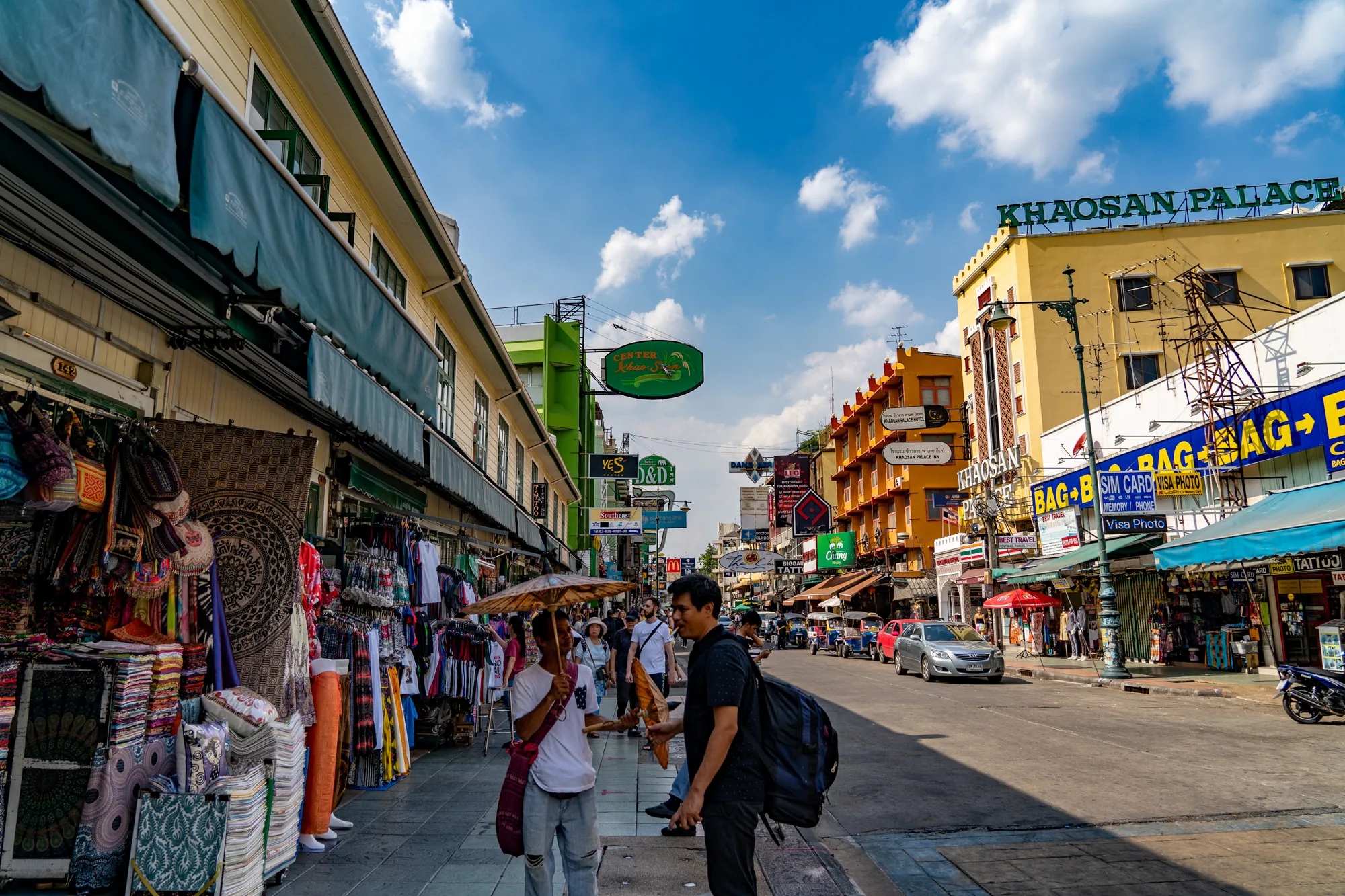 写真はタイのバンコクにあるカオサン通りを捉えたものです。晴れた日の昼間に撮影されており、青い空には白い雲が浮かんでいます。道の両側には建物が立ち並び、歩道には様々なお店が並んでいます。左側の歩道には、衣類、バッグ、雑貨などが所狭しと並べられ、観光客向けの露店が賑わっている様子がうかがえます。建物の壁には、カラフルな看板や広告が掲げられ、アジアの街並み特有の活気を感じさせます。  画面中央には道路が走り、車やトゥクトゥク（タイの三輪タクシー）が走行しています。歩道には人々が歩いており、傘を差している人もいます。画面手前には、リュックを背負った男性と、傘をさした男性が立っており、観光客の姿が見られます。全体的に賑やかで活気のある雰囲気であり、観光地らしいエネルギッシュな空気感が伝わってきます。Khaosan Palace Hotel や other hotelsの看板も確認できます。