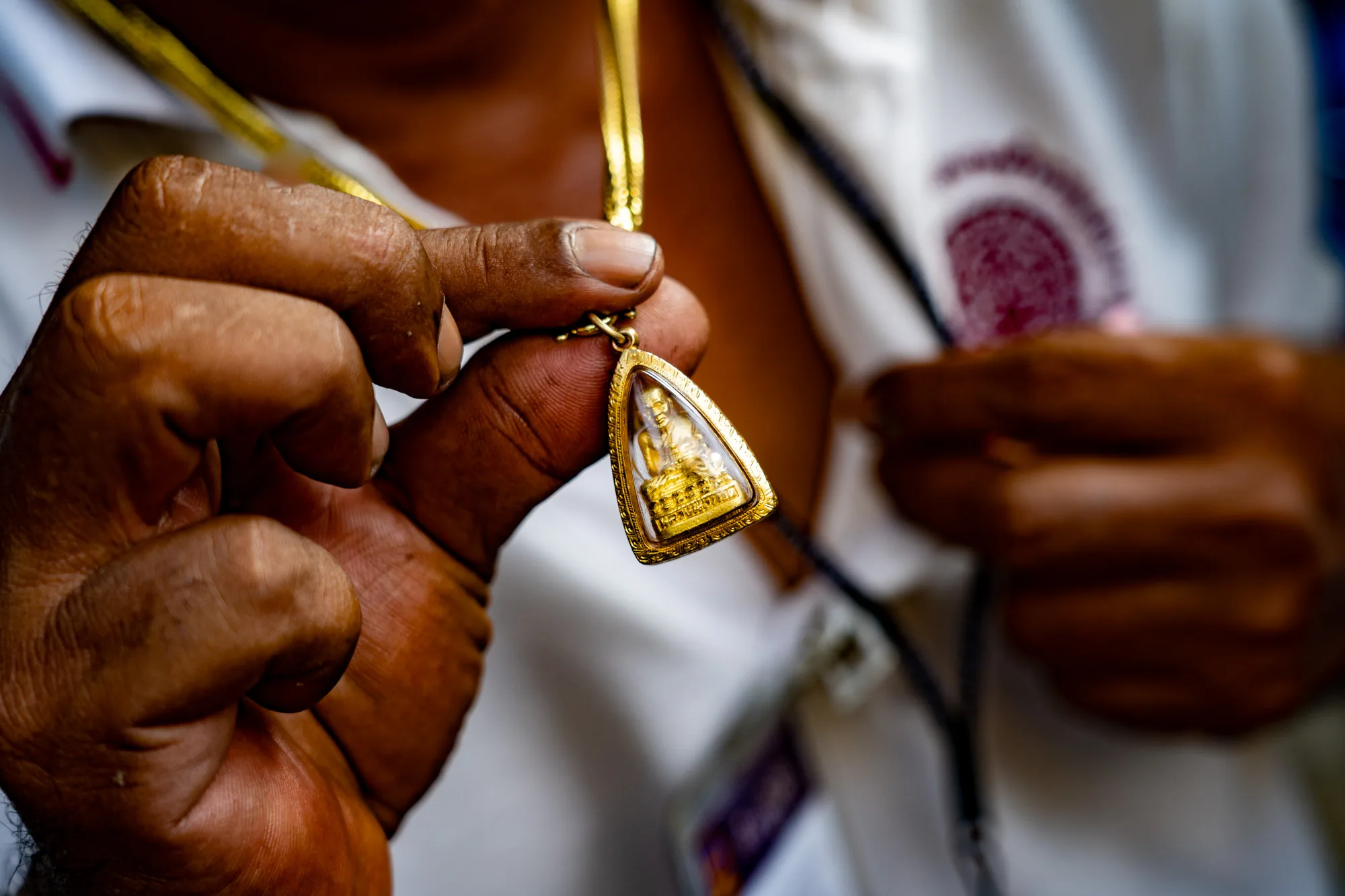 写真には、男性が手に持った仏像のお守りが写っています。男性の手は日焼けしており、指には細かな皺が見られます。お守りは金色のフレームで囲まれており、中には金色の仏像が納められています。仏像は台座の上に座っているように見えます。お守りは金のネックレスに通されています。男性は白いシャツを着ており、胸元が開いています。シャツには何か模様がプリントされていますが、ぼやけていて詳細は不明です。背景は暗く、被写体に焦点が当たっています。写真全体から、男性が大切にしているお守りであることが伝わってきます。
