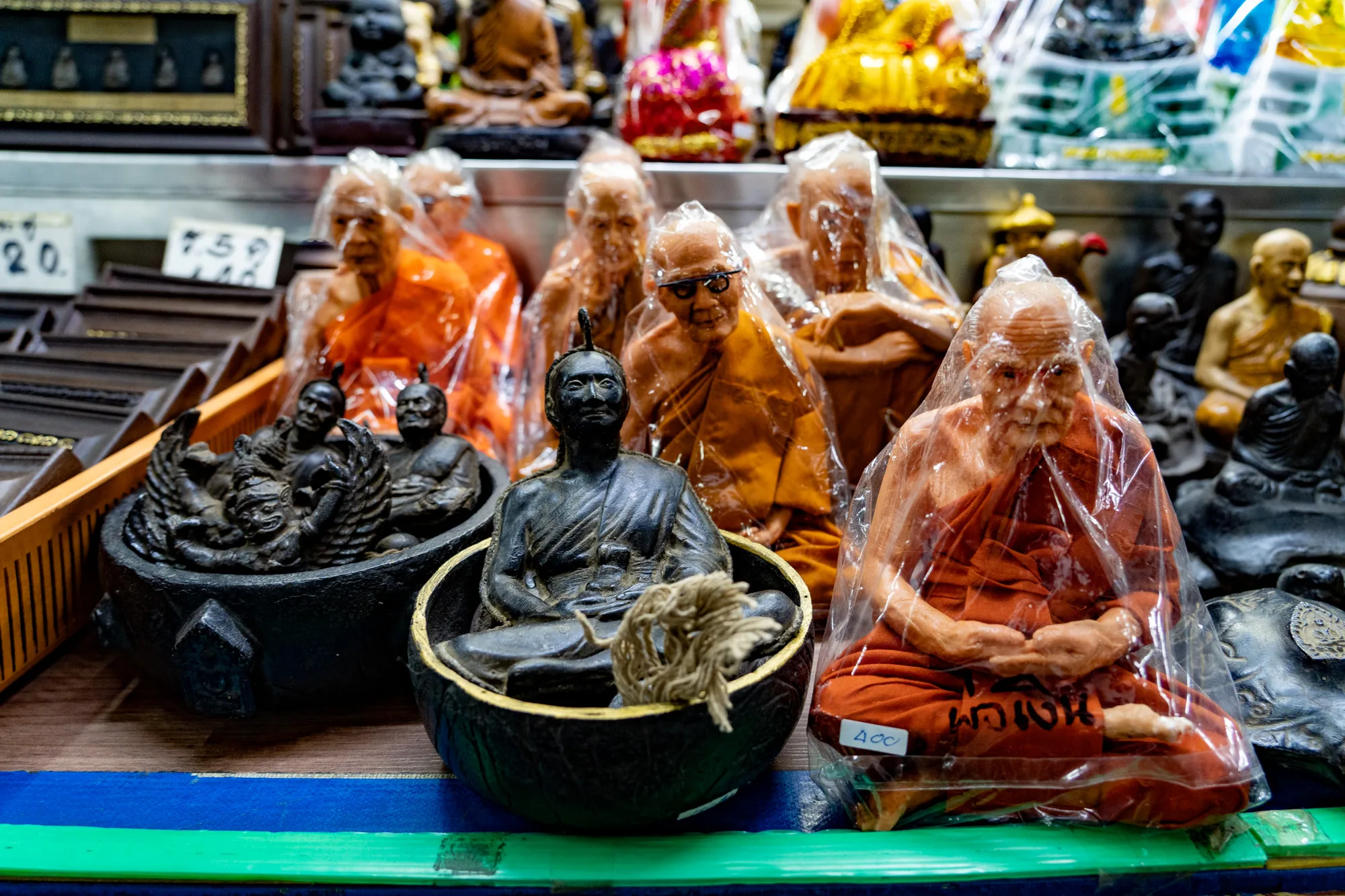 この写真は、タイのバンコクで撮影された仏像店の様子を捉えています。店内には、様々な種類の仏像や僧侶像が並べられています。手前には、黒色の容器に入った仏像や、オレンジ色の袈裟をまとった僧侶の像が、ビニール袋に入れられて陳列されています。僧侶の像の中には、メガネをかけたものもあり、表情豊かです。背景には、さらに多くの仏像が並んでおり、金色の装飾が施されたものや、色鮮やかな装飾が施されたものも見られます。全体的に、宗教的な雰囲気が漂い、タイの文化を感じさせる写真です。
