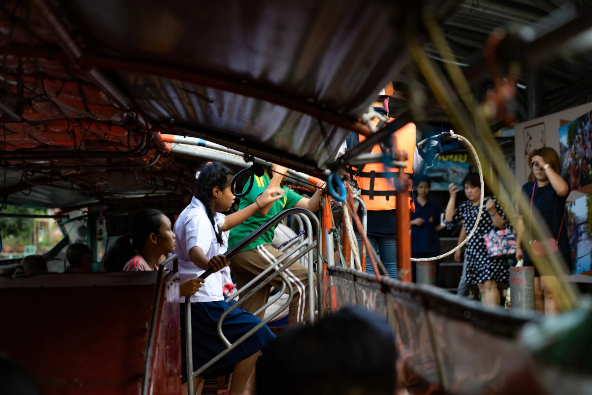 写真はタイのバンコクで撮影されたと思われる、水上ボートからの視点です。船内には制服を着た女子学生や、ライフジャケットを着用した人々がいます。学生たちは手すりにつかまり、何かを見ているようです。船の外には、ドット柄のワンピースを着た女性や、他の観光客らしき人たちが写っており、賑やかな雰囲気が伝わってきます。船の天井には救命胴衣が収納されており、ローカルな交通手段としてのボートの様子が伺えます。全体的に、活気のあるバンコクの風景を捉えたスナップショットです。