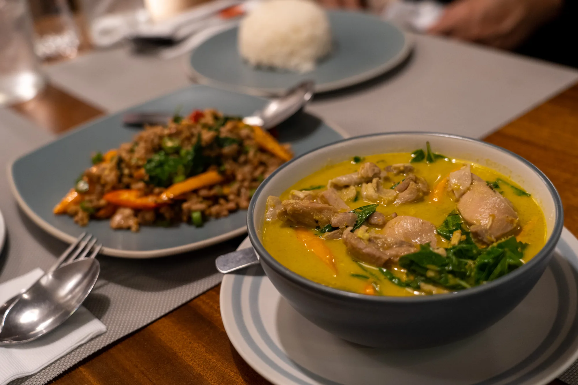 これはタイのバンコクで撮影された写真です。テーブルの上には、タイ料理の夕食が並べられています。手前には、鶏肉と野菜の入った黄色いカレーが入ったボウルがあります。ボウルはグレーの縁取りがされた白の皿に載っています。奥には、ひき肉と野菜を炒めた料理が青い皿に盛られています。さらに奥には、ご飯が青い皿に盛られています。テーブルには、フォークとスプーンが置かれ、食事を楽しむ準備ができています。全体的に、暖かく、食欲をそそる雰囲気です。