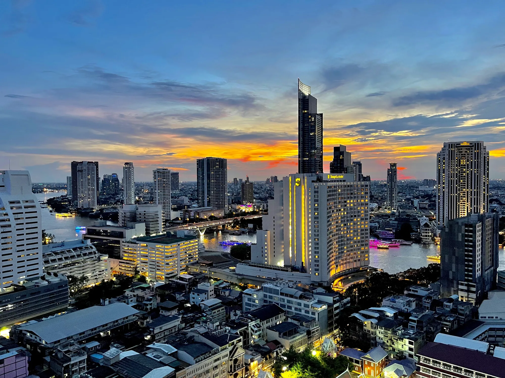 写真はタイのバンコクで撮影された夜景です。夕焼けの名残が残る空の下、チャオプラヤー川が蛇行し、その両岸には高層ビル群が立ち並んでいます。空は青からオレンジへとグラデーションを描き、雲がたなびいています。川面には、橋やボートの光が反射してきらめいています。  高層ビルは多様なデザインで、いくつかには明かりが灯っており、都市の活気を感じさせます。手前には低層の建物が密集したエリアがあり、その屋根もわずかにライトアップされています。全体的に、賑やかで美しいバンコクの夜の風景を捉えた写真です。