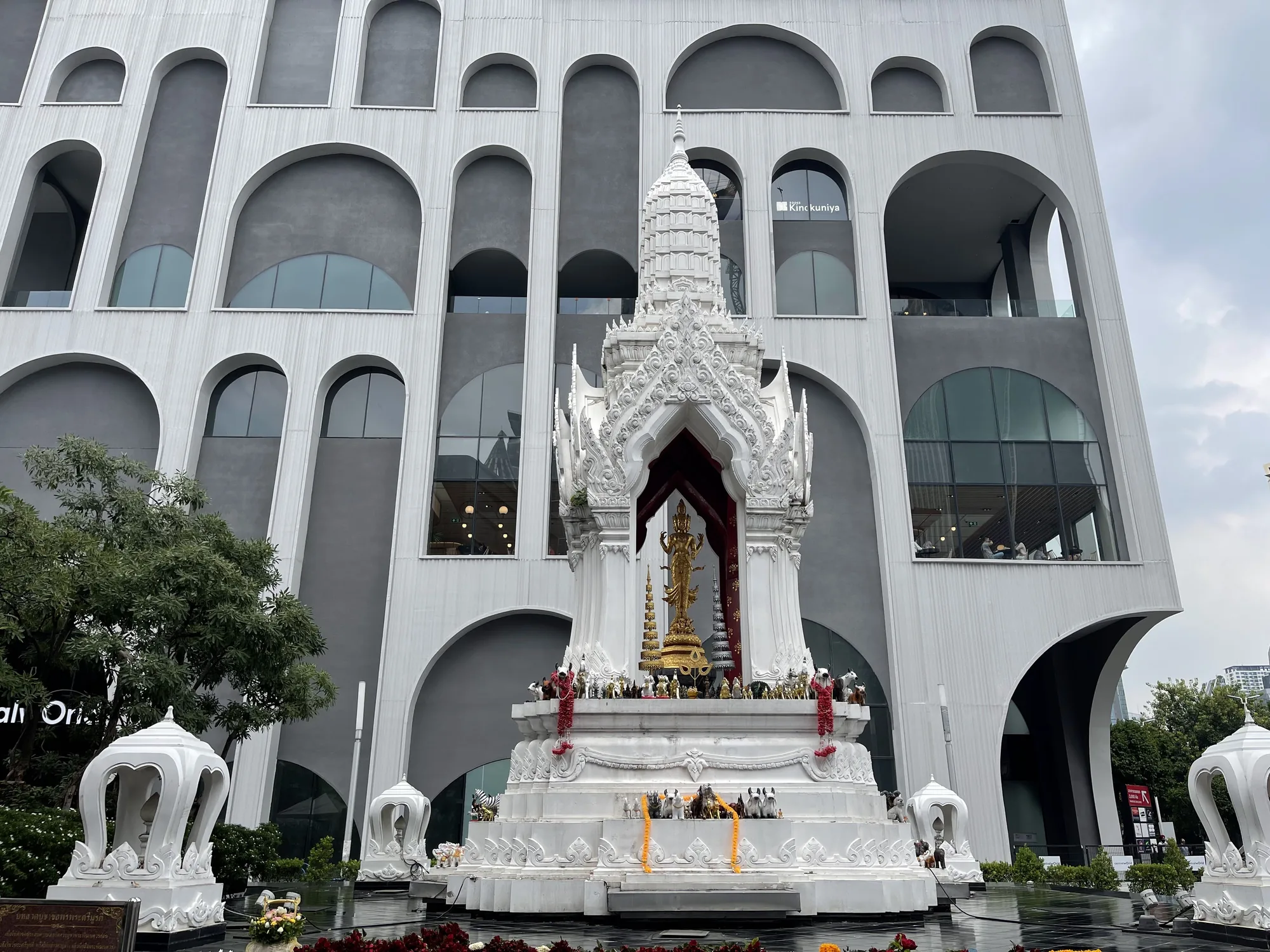 これはタイのバンコクで撮影された写真です。写真には、伝統的なタイ様式の白い祠が写っており、その中には金色の像が安置されています。祠は花や供物で飾られており、神聖な雰囲気を醸し出しています。祠の背景には、モダンなデザインの白い建物がそびえ立っています。建物は多数のアーチ状の窓が特徴的で、祠とのコントラストが印象的です。空は曇っており、全体的に落ち着いた雰囲気です。建物の窓には「Kinokuniya」の文字が見え、紀伊国屋書店であることがわかります。
