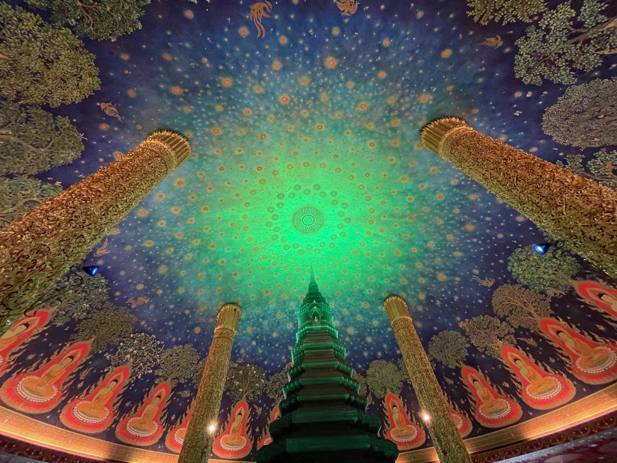 この写真は、タイのバンコクにある寺院内部の天井を見上げたものです。中央には緑色にライトアップされた仏塔がそびえ立ち、天井は深い青色を基調とし、中心部に向かって緑色へとグラデーションがかかっています。天井全体には、細かな星のような模様が無数に描かれており、まるで夜空のようです。さらに、天井の縁には、木や仏像が描かれた装飾的な壁画が施されています。写真には、金色の装飾が施された柱が複数本写っており、その豪華さが目を引きます。全体的に神秘的で荘厳な雰囲気を醸し出しており、タイの伝統的な寺院建築の美しさを感じさせます。