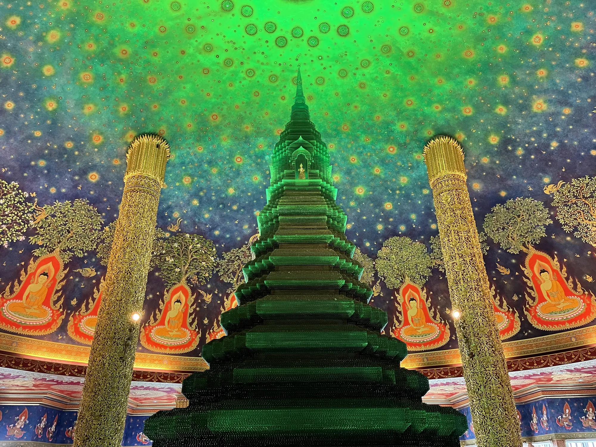 タイのバンコクで撮影された写真です。画面中央には、緑色にライトアップされたピラミッドのような仏塔がそびえ立っています。仏塔は多層構造で、各層には小さな窓のようなものが設けられています。最上部には小さな仏像が安置されているようです。  仏塔の両脇には、金色の装飾が施された柱が立っています。柱は細かく彫刻されており、複雑な模様が光を反射して輝いています。  背景の壁は、鮮やかな青色と緑色で彩られています。天井には、星空のような模様が描かれており、無数の星や花が散りばめられています。壁面には、仏陀の絵や、植物、動物などが描かれており、タイの伝統的な仏教美術の様式が見られます。  写真全体は、幻想的な雰囲気で満ちており、荘厳で神秘的な空間を表現しています。鮮やかな色彩と細部にまでこだわった装飾が、見る人を魅了するでしょう。