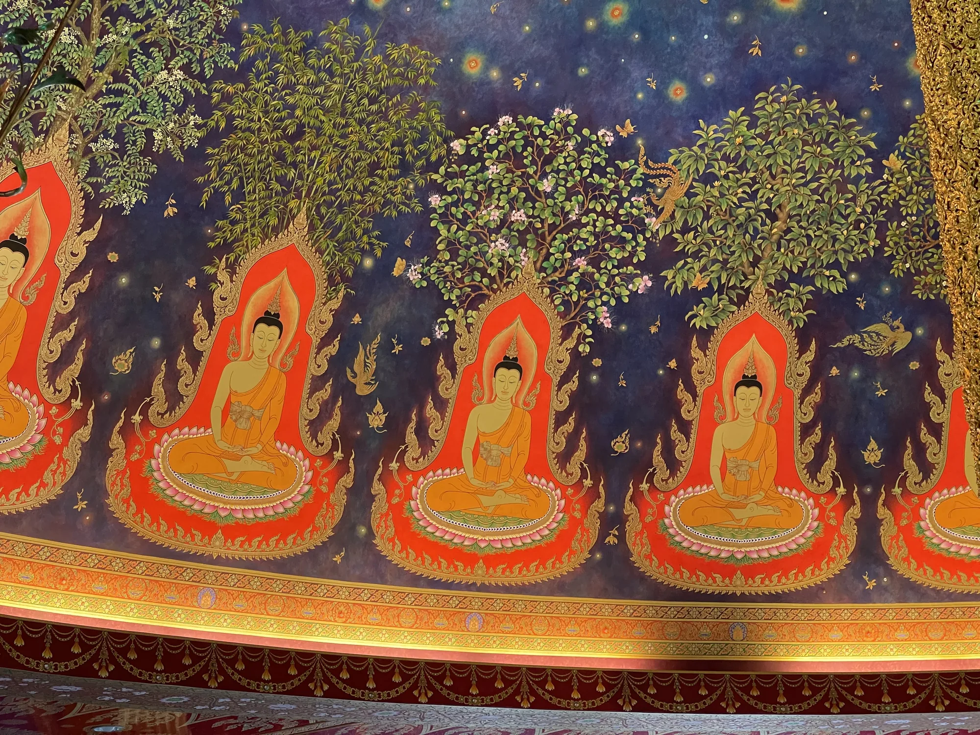 写真は、タイのバンコクにある寺院の天井を写したものです。天井には、複数の仏陀が描かれた美しい壁画が広がっています。仏陀は瞑想の姿勢で座っており、鮮やかなオレンジ色の衣を身につけています。仏陀の背景には、それぞれ異なる種類の樹木が描かれており、その上には星が散りばめられた深い青色の夜空が広がっています。夜空には、鳥や蝶、花などの装飾的な要素も描かれており、幻想的な雰囲気を醸し出しています。壁画の下部には、金色の装飾が施された帯が描かれており、壁画全体の豪華さを引き立てています。全体的に、この写真はタイの寺院の荘厳さと美しさを伝える印象的な一枚です。