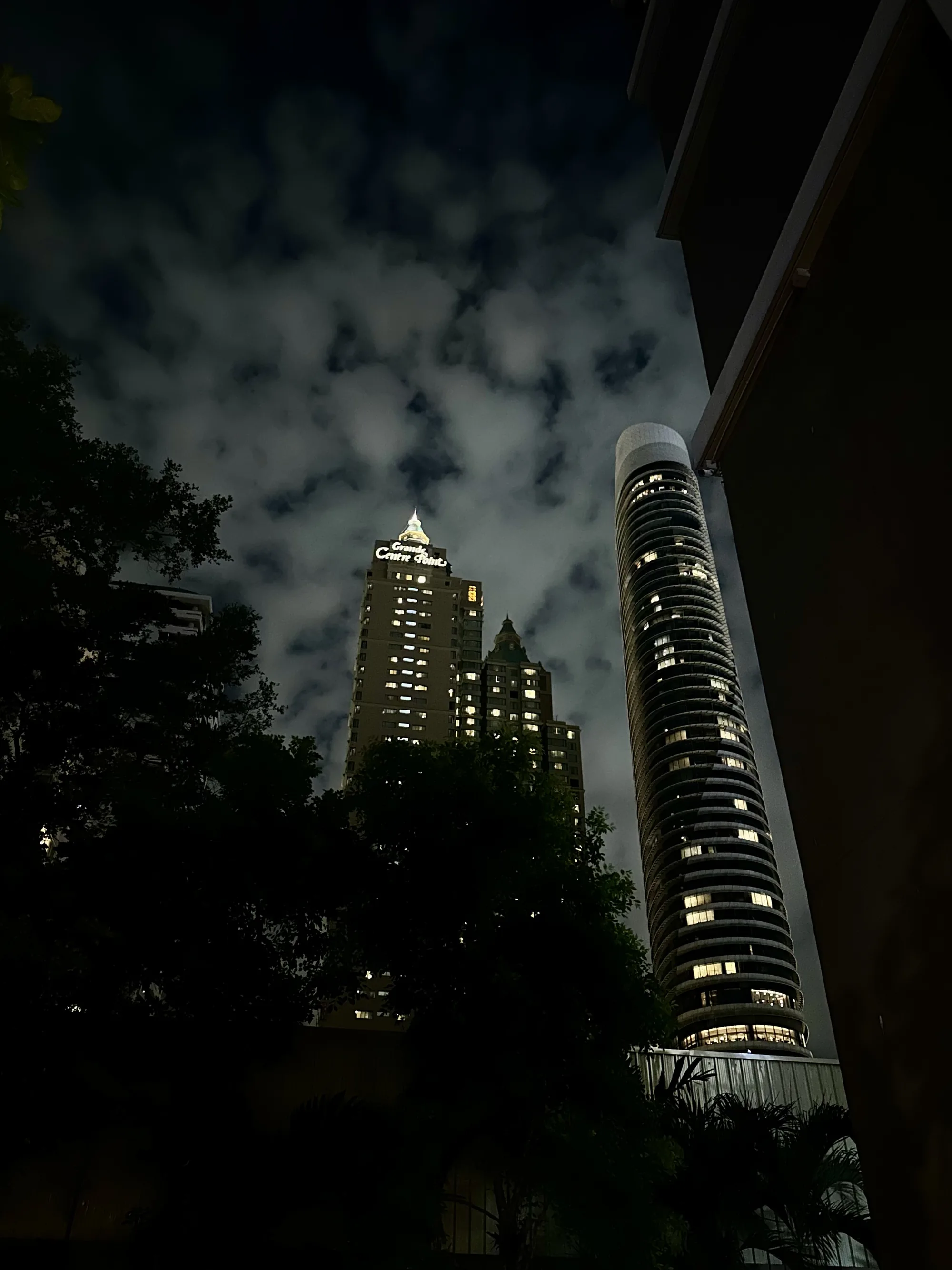 タイのバンコクで撮影された夜景の写真です。画面全体は暗く、夜のしっとりとした雰囲気が漂っています。空には雲が広がり、その間から覗く夜空が深みを感じさせます。  写真には二つの高層ビルが写っており、それぞれ異なる形状をしています。左側のビルは「Grande Centre Point」と書かれた看板を掲げており、その文字が光を放っています。ビルの窓からは光が漏れ、人々の生活の息吹を感じさせます。右側のビルは円筒形で、こちらも同様に窓から光が漏れています。  手前には街路樹のシルエットが重なり、都会の風景に自然の要素を加えています。写真全体が、暗闇と光のコントラストによって、都会の夜の表情を豊かに表現しています。
