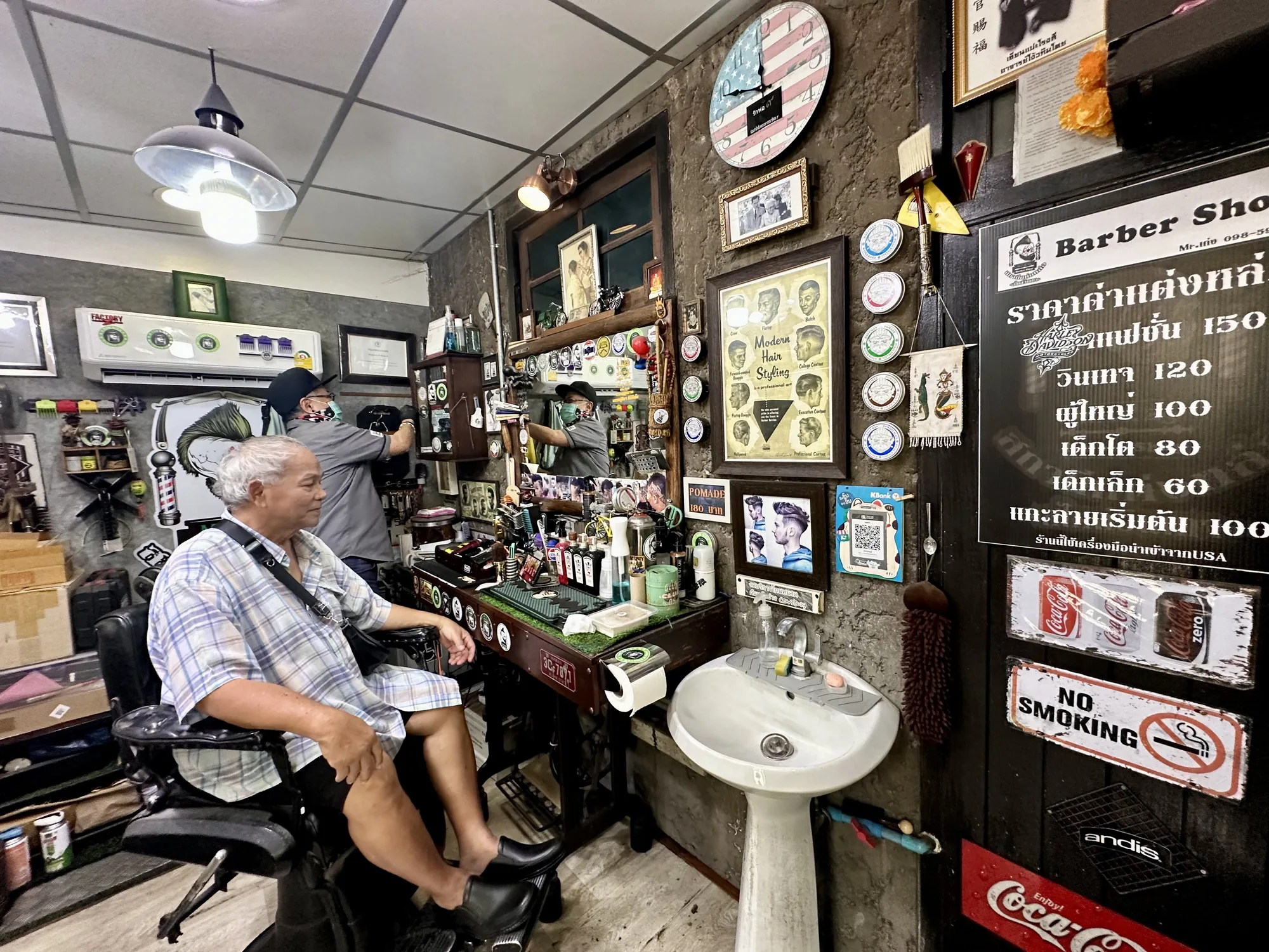 写真はタイのバンコクにあると思われる、レトロな雰囲気の理髪店を捉えています。店内の様子は、壁に飾られた古いポスターや装飾品から、昔ながらのスタイルを保っていることが伺えます。店内には理髪椅子に座る男性客と、マスクをした理容師の姿が見えます。理容師は、鏡に映る自分の姿を見ながら、作業をしているようです。壁にはアメリカ国旗を模した時計、昔のヘアスタイルを紹介するポスター、そしてタイ語で書かれた料金表などが飾られています。洗面台や、コカ・コーラの古い看板などもあり、ノスタルジックな雰囲気を醸し出しています。全体的に、歴史を感じさせる、趣のある理髪店の様子が伝わってくる写真です。