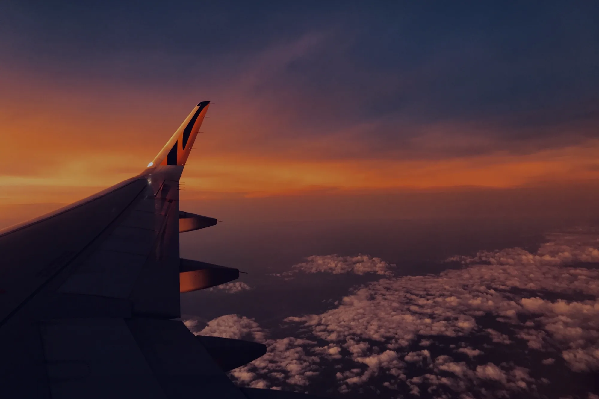 写真は、飛行機の窓から見た風景を捉えたものです。機体の翼の一部が画面左側に大きく写っており、その向こうには、夕焼けに染まる空と雲海が広がっています。夕焼けの色は、オレンジ色から紫色へとグラデーションを描いており、空全体を幻想的な雰囲気に包み込んでいます。眼下には、綿菓子のような白い雲が広がり、その上に夕焼けの光がほんのりと反射しています。この写真からは、空の旅の解放感と、夕焼けの美しさによる感動が伝わってきます。撮影場所がタイのバンコクとのことなので、熱帯の空ならではの、力強く鮮やかな色彩が印象的です。