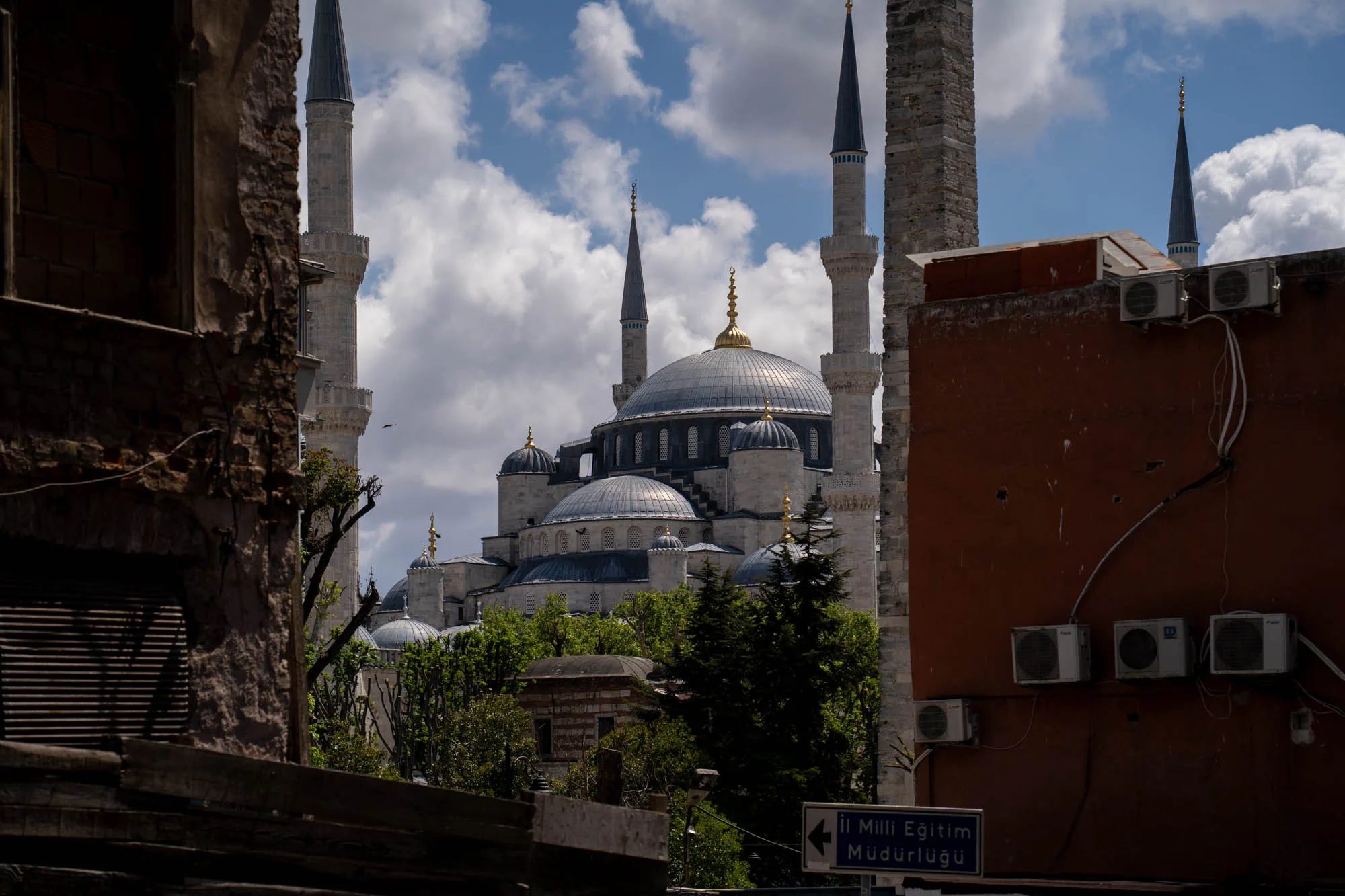 写真はトルコのイスタンブールで撮影されたもので、中央には有名なブルーモスク（スルタンアフメト・モスク）が堂々とそびえ立っています。モスクは巨大なドームと、高くそびえ立つミナレット（尖塔）が特徴的で、空の青さと白い雲を背景に美しいシルエットを描いています。手前には、歴史を感じさせる古い建物の一部が左右に配置され、モスクへの視線を額縁のように囲んでいます。左側の建物はレンガ造りで、ところどころ崩れかけており、歴史の重みを感じさせます。右側の建物は茶色で、壁には複数のエアコンの室外機が取り付けられており、生活感があります。また、"Il Milli Eğitim Müdürlüğü"と書かれた青い標識が見えます。全体的に、歴史的な建造物と現代の都市の風景が共存している様子が写し出されています。