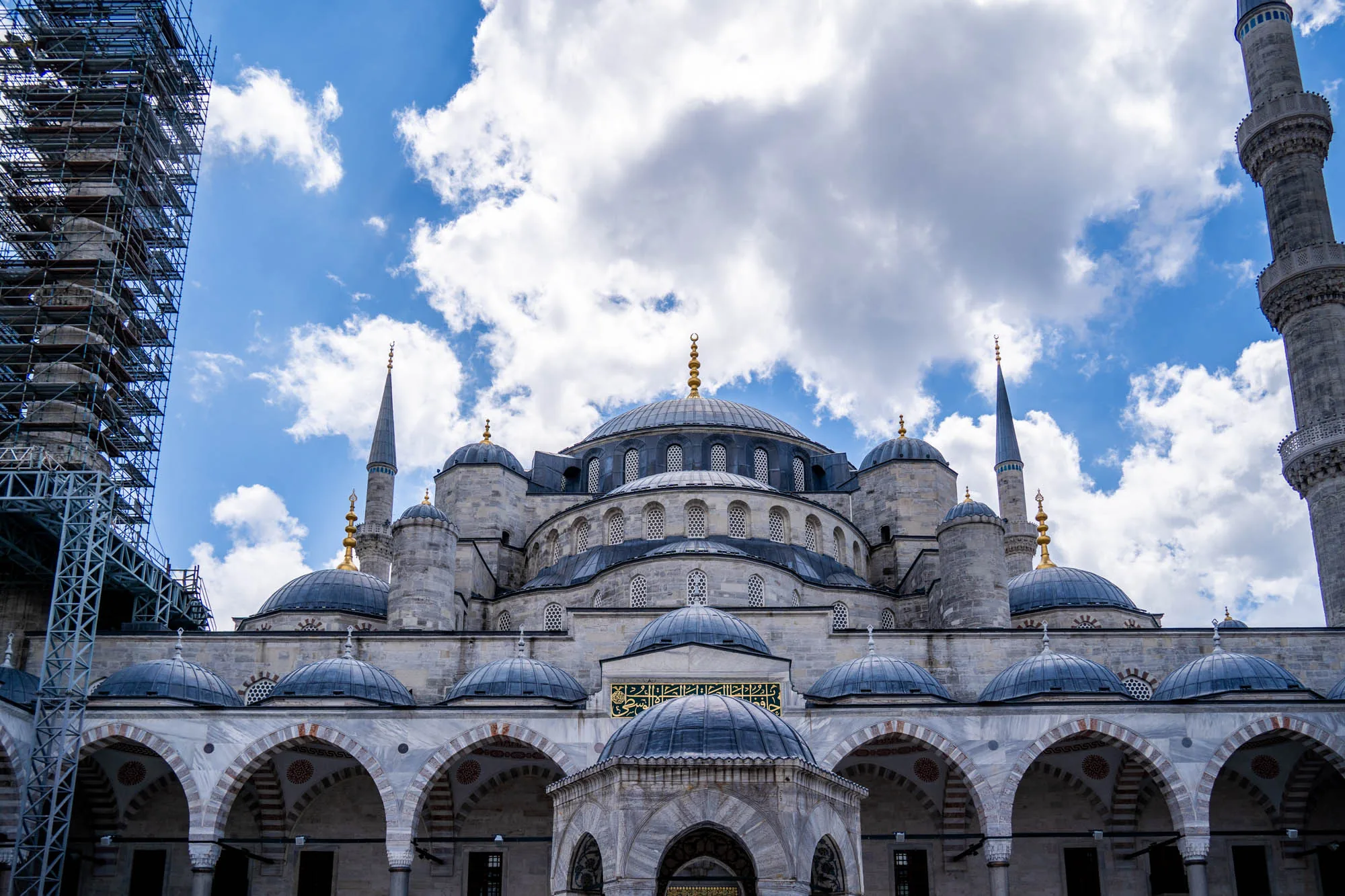 写真には、トルコのイスタンブールにある壮大なモスクが写っています。青い空と白い雲を背景に、モスクの複雑な構造が際立っています。複数のドームが重なり合い、ミナレットがそびえ立っています。一部のミナレットには修復用の足場が組まれており、歴史的建造物の維持活動が行われていることがわかります。建物の下部には美しいアーチが連なり、その装飾的な模様が細部まで表現されています。全体的に、荘厳で落ち着いた雰囲気が感じられる写真です。