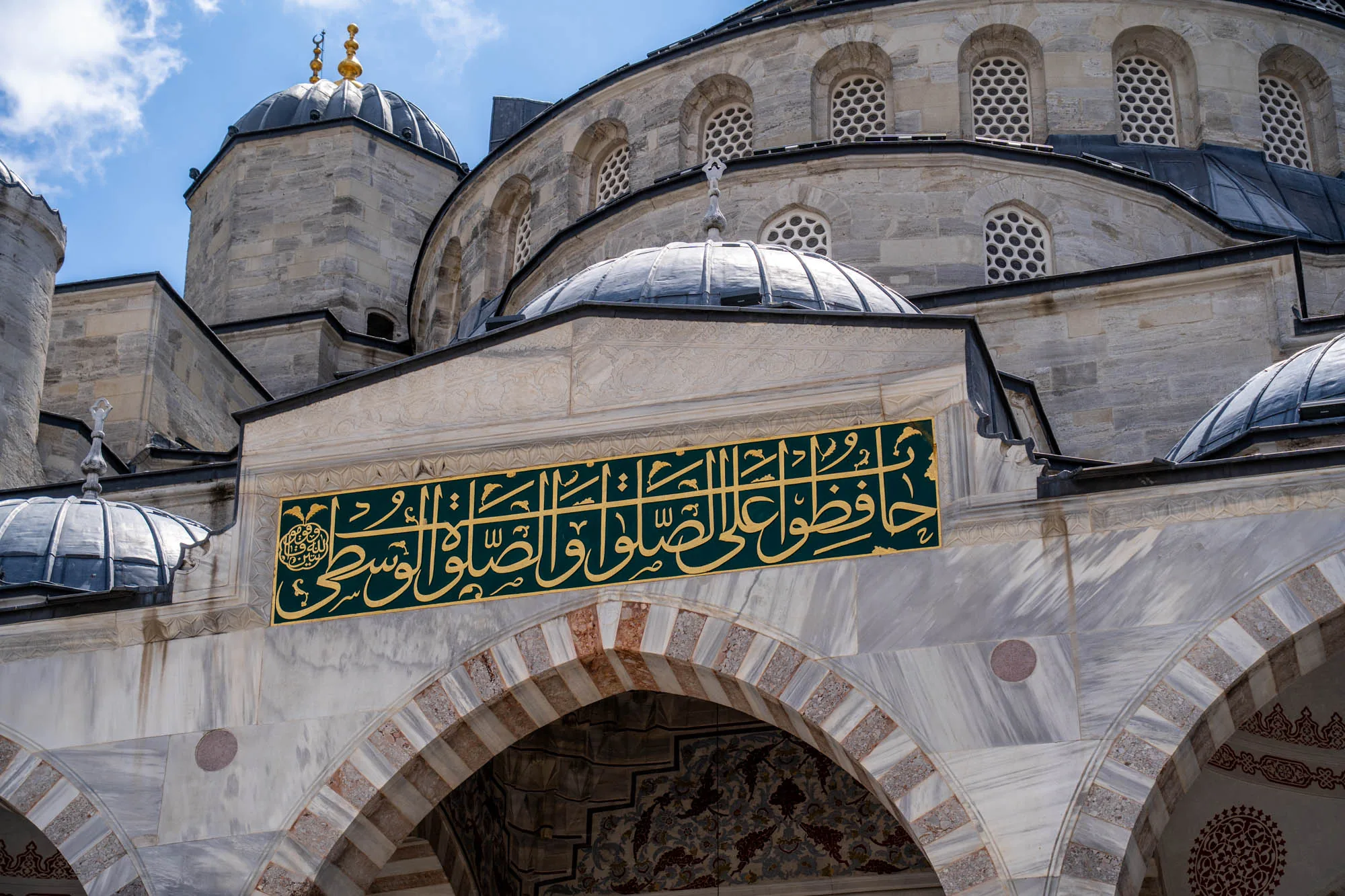 この写真は、トルコのイスタンブールにあるモスクの一部を捉えたものです。画像には、石造りの壁面と複数のドーム状の屋根が写っています。壁面は白とグレーの大理石で構成されており、美しい模様が施されています。壁の上部には、緑色の背景に金色のカリグラフィー（アラビア文字）が描かれた装飾的なパネルがあります。  モスクの屋根は複数のドームで構成されており、それぞれ異なるサイズと高さを持っています。ドームは鉛色の金属で覆われており、一部のドームの頂点には金色の装飾が施されています。ドームの周りには、小さく丸い窓が規則的に配置されています。  写真全体から、モスクの壮大さと精巧な建築様式が伝わってきます。青い空と白い雲が背景に写っており、建物の荘厳さを引き立てています。全体的に、落ち着いた雰囲気で、細部まで丁寧に作られたイスラム建築の美しさを感じさせる一枚です。