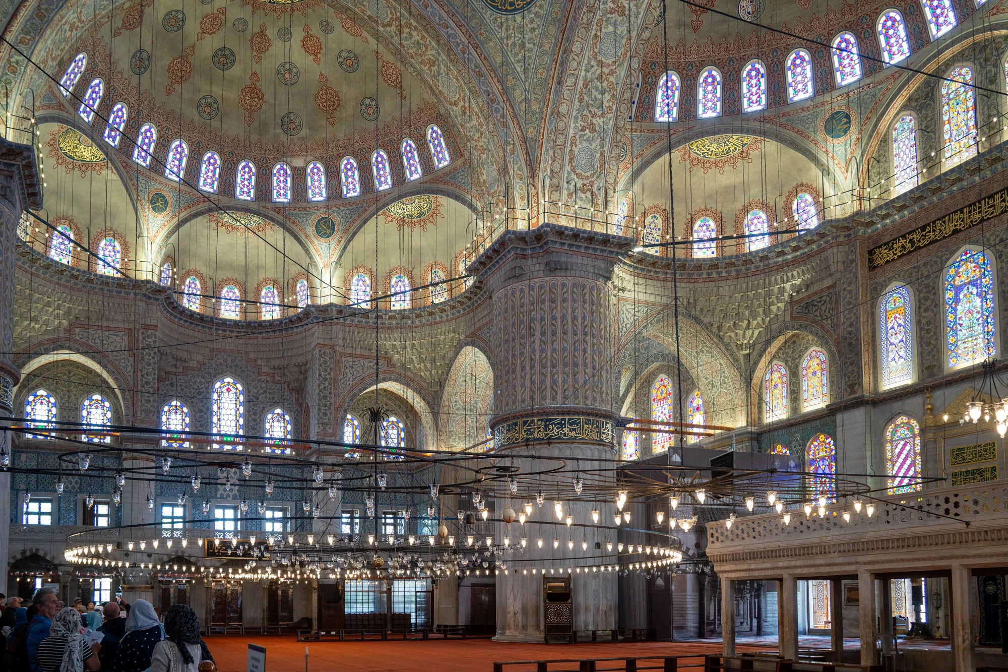 写真はトルコのイスタンブールにある有名な青モスクの内部を捉えています。巨大なドーム型の天井は精緻な模様で覆われ、赤、青、緑などの色鮮やかなステンドグラスの窓が並んでいます。天井を支える巨大な柱もまた、美しい装飾が施されています。床は赤い絨毯で覆われ、シャンデリアがいくつも吊り下げられています。シャンデリアの明かりが空間全体を照らし、荘厳で神秘的な雰囲気を醸し出しています。写真の下部には、観光客と思われる人々の姿が小さく写っており、この場所の大きさを際立たせています。モスクの内部は広大で、細部までこだわり抜かれた装飾が施されており、見る者を圧倒する美しさです。