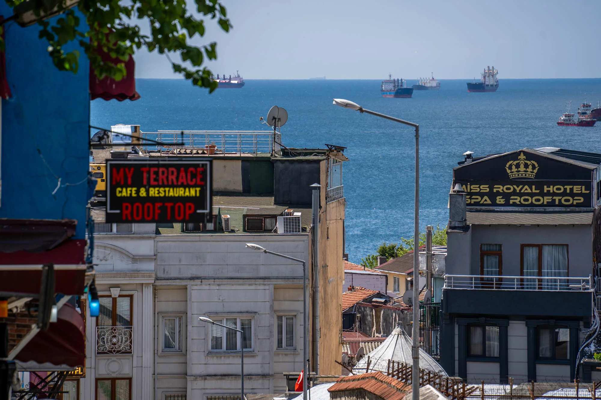 この写真は、トルコのイスタンブールで撮影された街並みの風景です。手前に複数の建物が立ち並び、その奥には広大な海が広がっています。建物はそれぞれ異なる高さとデザインを持ち、屋上にはレストランやホテルの看板が見えます。海には複数の大型船が停泊しており、遠くまで続く水平線が広がっています。青い空と海のコントラストが美しく、都市の喧騒と海の静けさが共存する風景が印象的です。