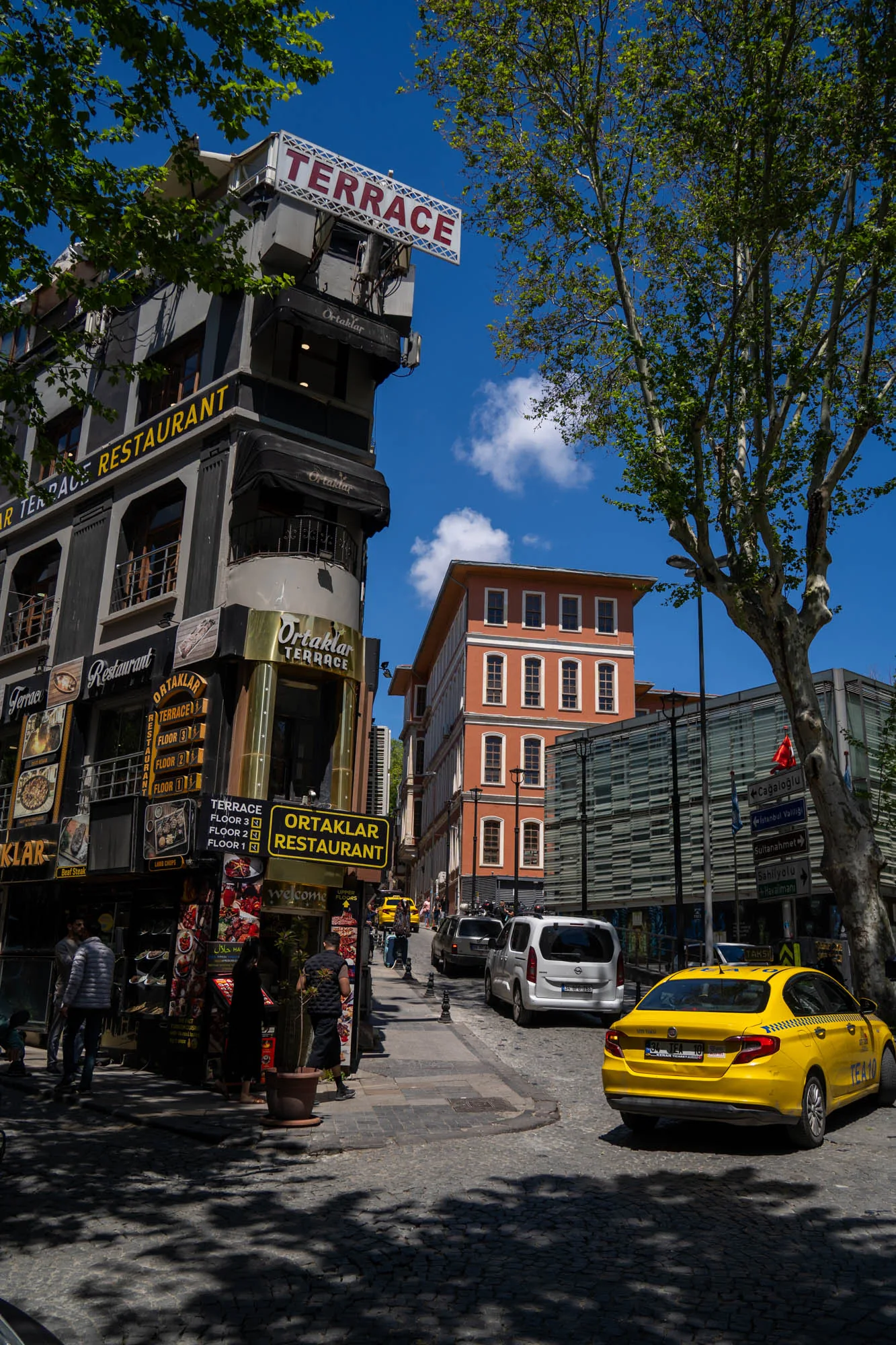 写真はトルコのイスタンブールで撮影された、賑やかな街の様子を捉えています。石畳の道路沿いには、複数の階を持つレストラン「Ortaklar Terrace Restaurant」がそびえ立ち、その看板が目を引きます。レストランの壁面には、提供される料理の写真が多数飾られており、食欲をそそります。  道路にはイエローキャブが停車しており、歩道には人々が行き交っています。空は晴れ渡り、青空が広がっています。画面上部には緑豊かな木々が写り込んでおり、都市の中にも自然が感じられます。奥にはオレンジ色の建物も見え、歴史的な街並みが垣間見えます。全体的に、活気があり、観光地らしい雰囲気が漂っています。