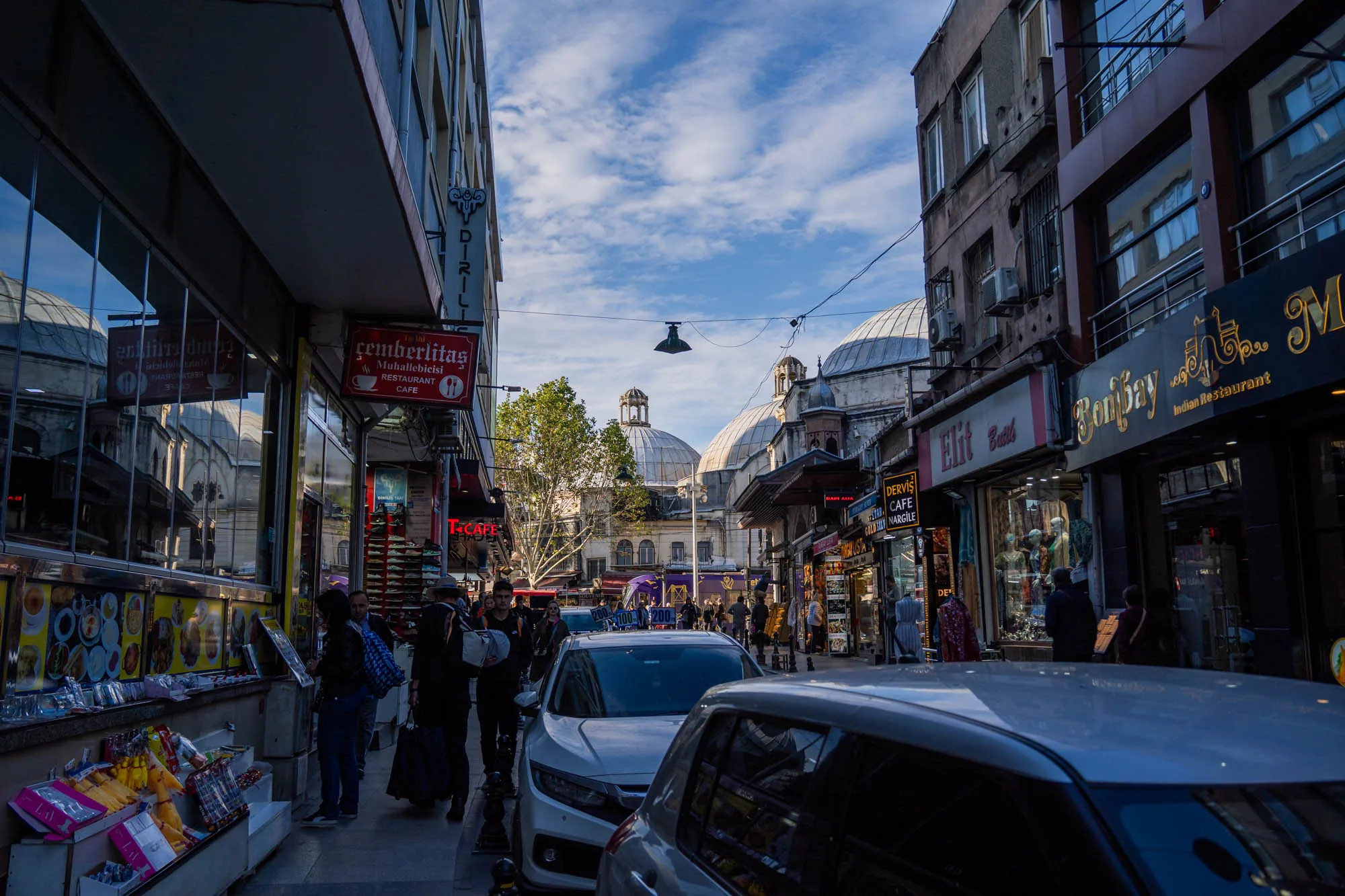 写真はトルコのイスタンブールで撮影された街並みを捉えています。通りは建物が両側に立ち並び、多くの人々が行き交っています。画面奥には特徴的なドーム型の建物が見え、イスタンブールらしい景観を作り出しています。手前には車が数台駐車しており、歩道には観光客らしき人々が歩いています。通り沿いにはレストランやカフェ、土産物店などが並んでおり、賑やかな雰囲気です。「çemberlitaş Muhallebicisi」と書かれた看板が見えることから、チェンベルリタシュ地区であると推測できます。全体的に、歴史的な建造物と現代の都市生活が混ざり合ったイスタンブールの魅力が感じられる写真です。
