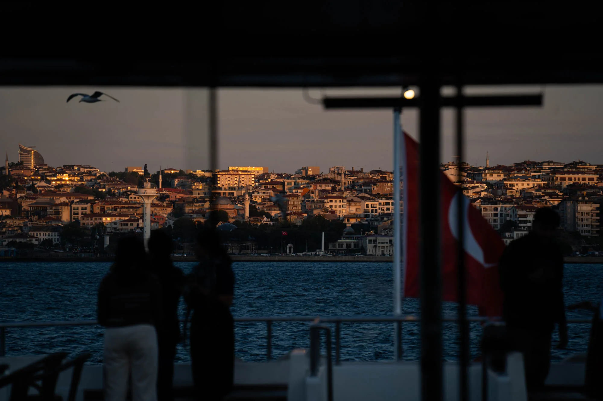 写真はトルコのイスタンブールで撮影されたもので、夕暮れ時の風景を捉えています。手前にはフェリーの船内の一部が写っており、乗客のシルエットが見えます。船内からは、イスタンブールの街並みが広がっており、建物が密集して立ち並ぶ様子が伺えます。夕日に照らされた建物は、オレンジ色に輝き、美しいコントラストを生み出しています。海には穏やかな波が立ち、遠くにはカモメが飛んでいるのが見えます。トルコの国旗が掲げられており、この場所がトルコであることを示しています。全体的に落ち着いた雰囲気で、夕暮れの静けさとイスタンブールの街並みの美しさが感じられる写真です。