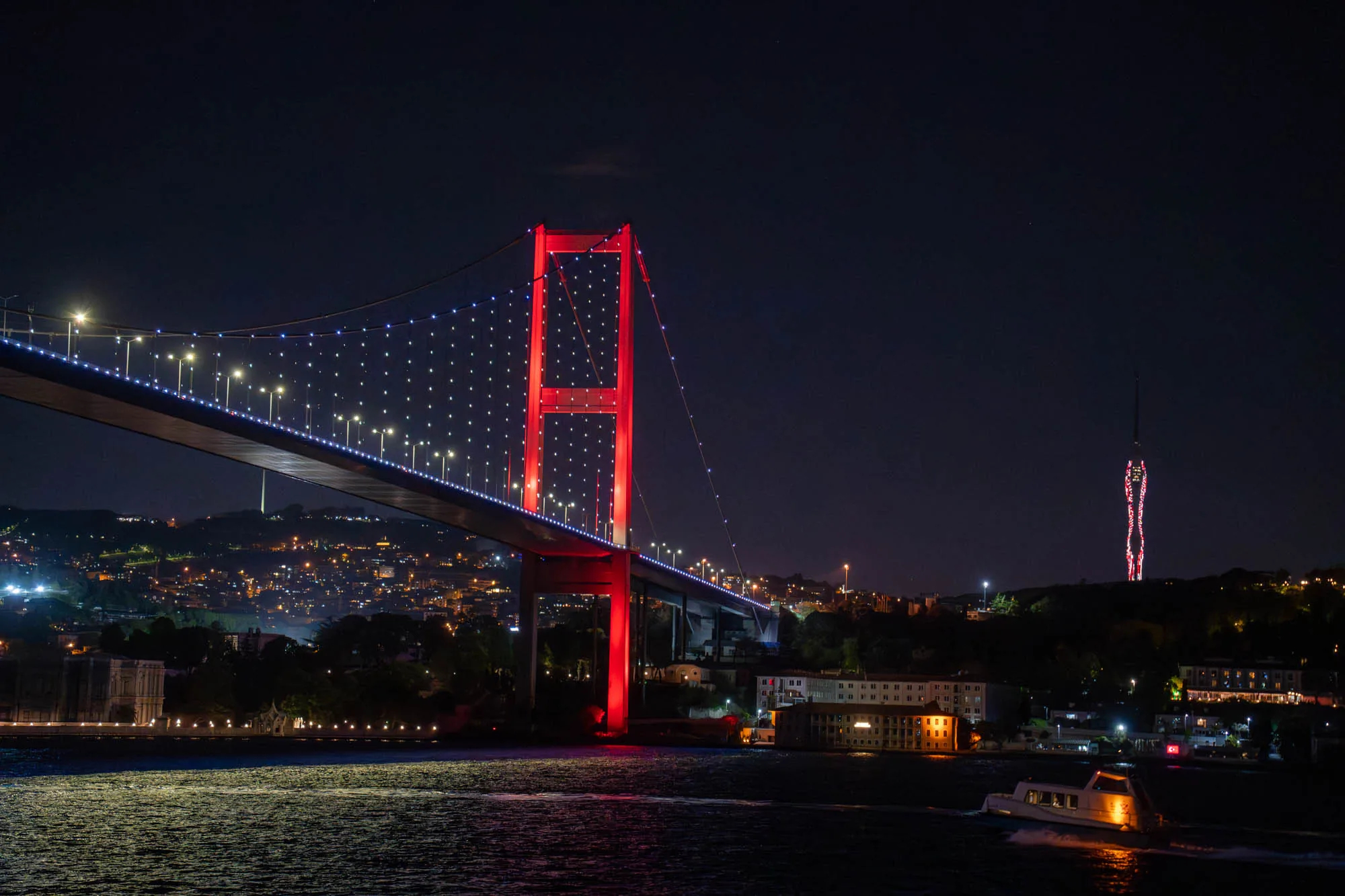 これはトルコのイスタンブールで撮影された夜景の写真です。主要な被写体は、ライトアップされたボスポラス海峡に架かる橋です。橋の主塔は鮮やかな赤色に照らされ、橋桁には小さな白いライトが連なり、夜空に浮かび上がるように輝いています。遠景には、市街地の明かりが広がり、丘陵に沿って建物が立ち並んでいるのが見えます。右奥には、同じく赤くライトアップされた高い塔がそびえ立っています。手前の水面は暗く、光を反射してきらめいています。小さなボートが一隻、ライトを点けて航行しており、夜の海に航跡を描いています。全体的に、夜のイスタンブールの美しい景観を捉えた、幻想的な雰囲気の写真です。