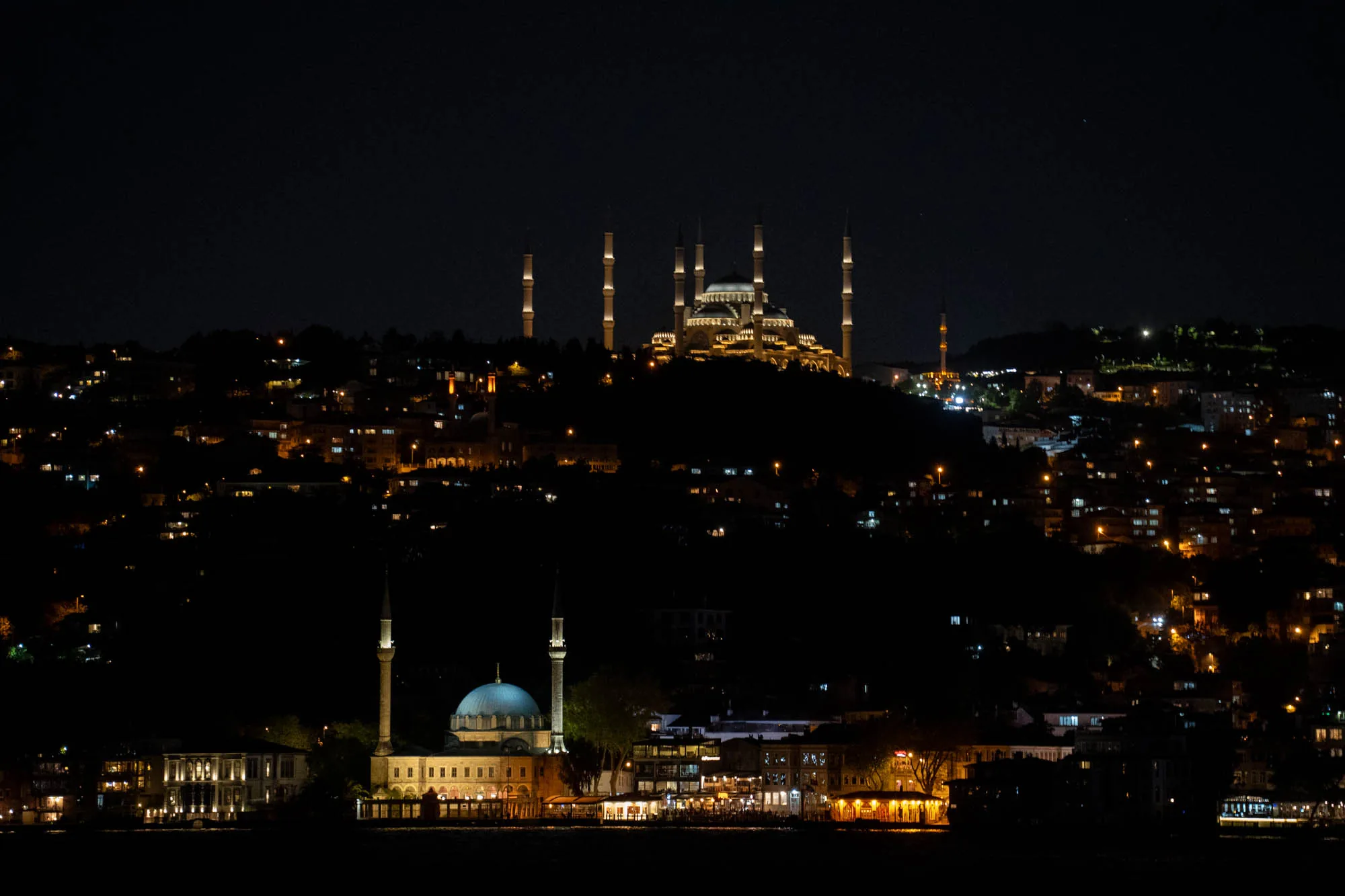 写真は、トルコのイスタンブールで撮影された夜景です。手前に海が広がり、その向こうには建ち並ぶ建物群が見えます。特に印象的なのは、ライトアップされたモスクです。丘の上にそびえ立つ大きなモスクは、複数のミナレット（尖塔）を持ち、その美しいシルエットが夜空に浮かび上がっています。手前にもモスクがあり、こちらもライトアップされていて存在感があります。周囲の建物や道路もライトアップされており、都市の活気が感じられます。夜空は暗く、建物の光が際立っています。全体的に落ち着いた雰囲気で、イスタンブールの美しい夜の風景を捉えた写真です。