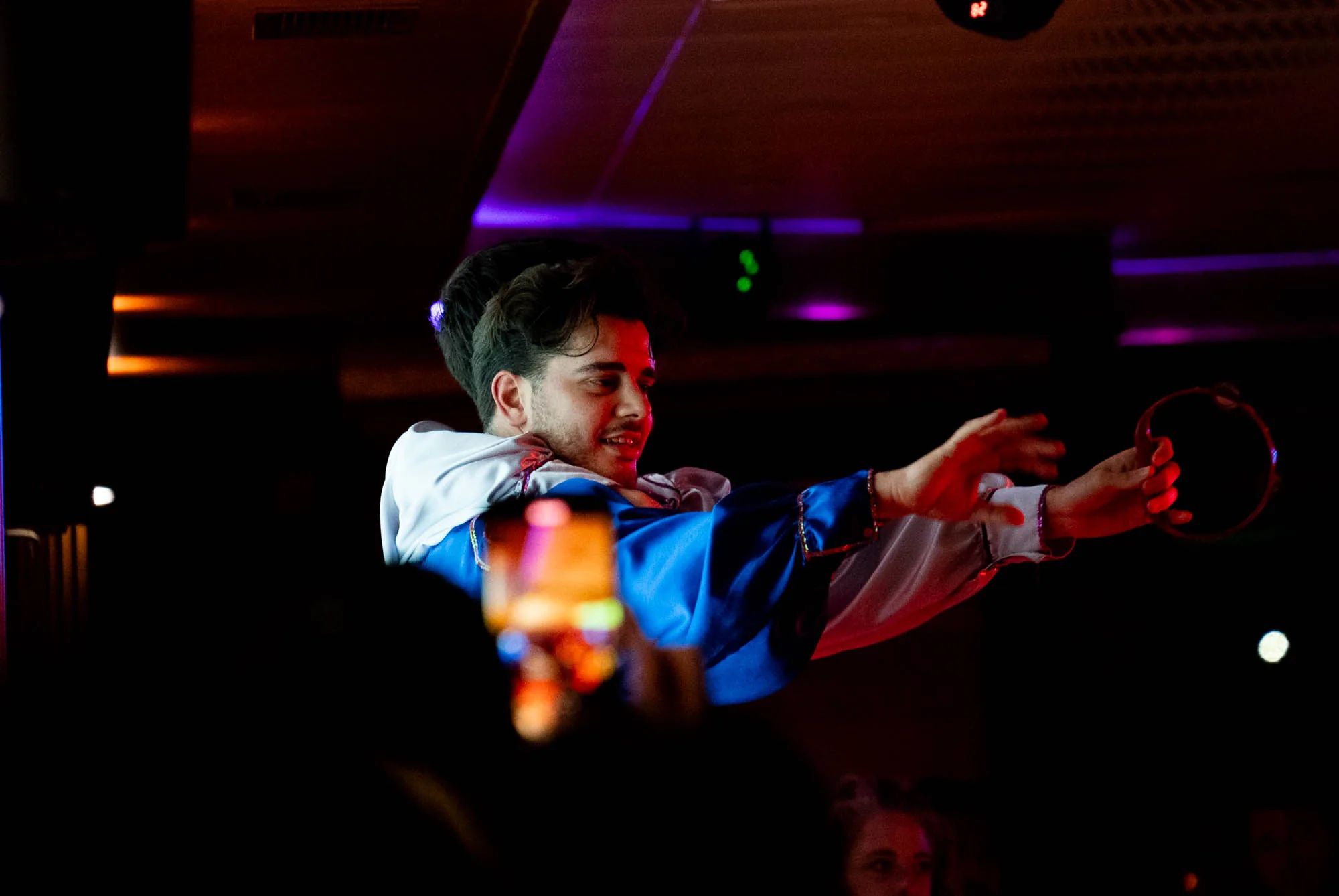 この写真は、イスタンブールで撮影されたと思われる、タンバリンを演奏する男性の姿を捉えています。薄暗い照明の下、男性は青と白の伝統的な衣装を身に着け、笑顔でタンバリンを叩いています。観客が彼のパフォーマンスをスマートフォンで撮影している様子も写っており、ライブ感あふれる雰囲気です。天井の照明が紫やオレンジ色に光り、背景を彩っています。