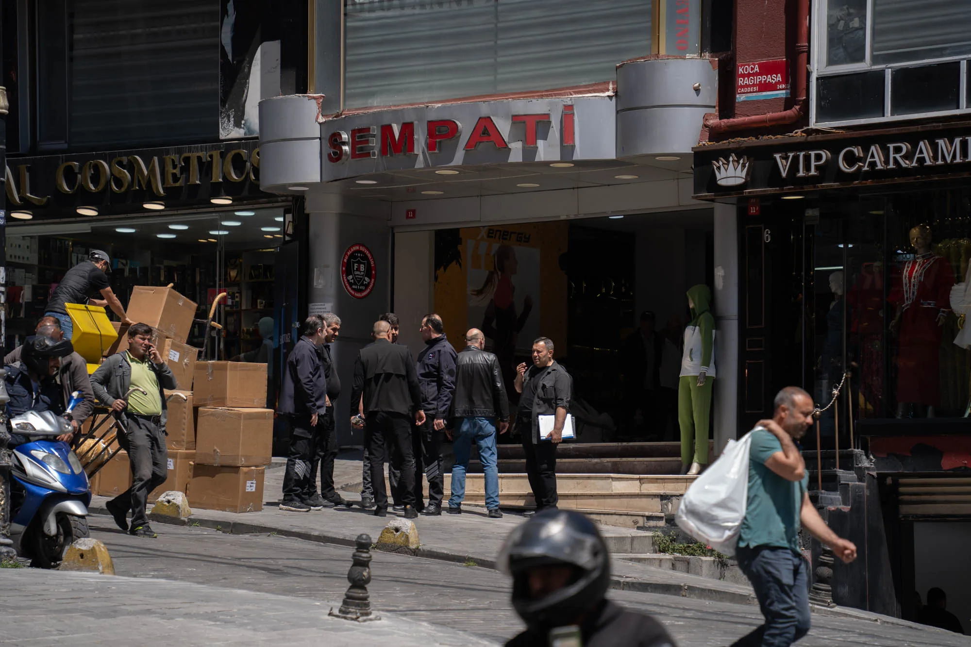 写真はトルコのイスタンブールで撮影された街並みをとらえています。中央には「SEMPATI」と書かれた看板の建物があり、その前には数人の男性が集まって話をしています。男性たちは黒いジャケットやシャツを着ている人が多く、ビジネスマンのような印象です。  左側には「AL COSMETICS」という化粧品店の看板が見え、配達員がスクーターで荷物を運んでいる様子が写っています。右側には「VIP CARAMI」という店があり、マネキンが飾られています。通りには歩行者もおり、生活感のある風景です。写真全体は明るく、日中の様子であることがわかります。道標には「KOCA RAGIPPAŞA CADDESI」と書かれており、通りの名前を示しています。全体として、賑やかなイスタンブールの街の日常を切り取った写真と言えるでしょう。