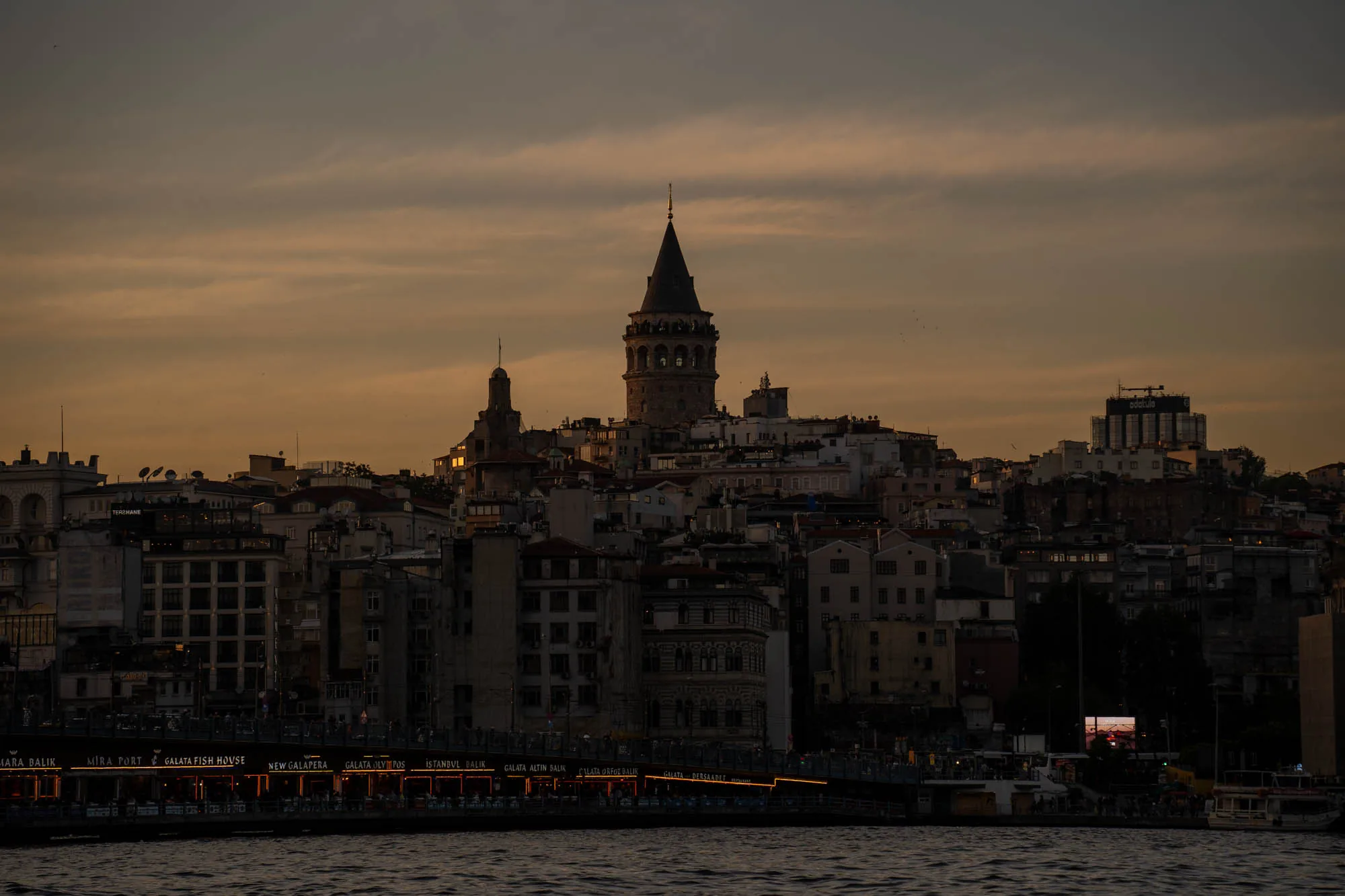 この写真はトルコのイスタンブールで撮影されたものです。夕暮れ時の空は、淡いオレンジ色と灰色が混ざり合い、穏やかな雰囲気を醸し出しています。手前には水面が広がり、穏やかな波が立っています。  水辺には、カラフルなライトで装飾されたレストランやショップが並んでいます。これらの灯りが水面に反射し、美しい光の模様を描き出しています。店の看板には「MARA BALIK」「MIRA PORT」「GALATA FISH HOUSE」「NEW GALAPERA」「GALATA OLYMPOS」「ISTANBUL DALIK」などの文字が見えます。  背景には、イスタンブールの街並みが広がっています。古い建物が密集しており、その中にそびえ立つガラタ塔が印象的です。ガラタ塔は、街のランドマークとして夕日に照らされ、シルエットのように浮かび上がっています。