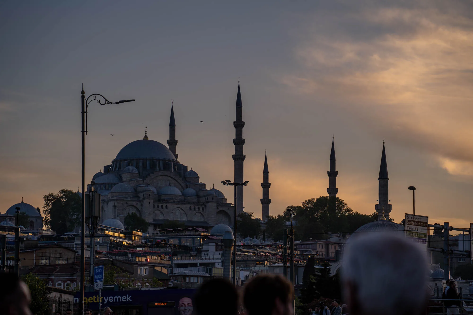 イスタンブールで撮影された、モスクのシルエットが印象的な夕景の写真です。空はオレンジとグレーが混ざり合い、夕焼けの温かさと夜の訪れを感じさせます。手前には人々の頭部がシルエットで写っており、観光客が多い場所であることが伺えます。遠景には、複数のミナレット（尖塔）を持つ巨大なモスクが見えます。モスクのドームや壁の一部が夕日に照らされ、金色に輝いています。モスクの周りには建物が密集しており、その上にモスクがそびえ立つ様子が印象的です。電柱や標識なども写っており、街の様子を伝えています。全体的に落ち着いた色調で、静かで美しい雰囲気を醸し出しています。
