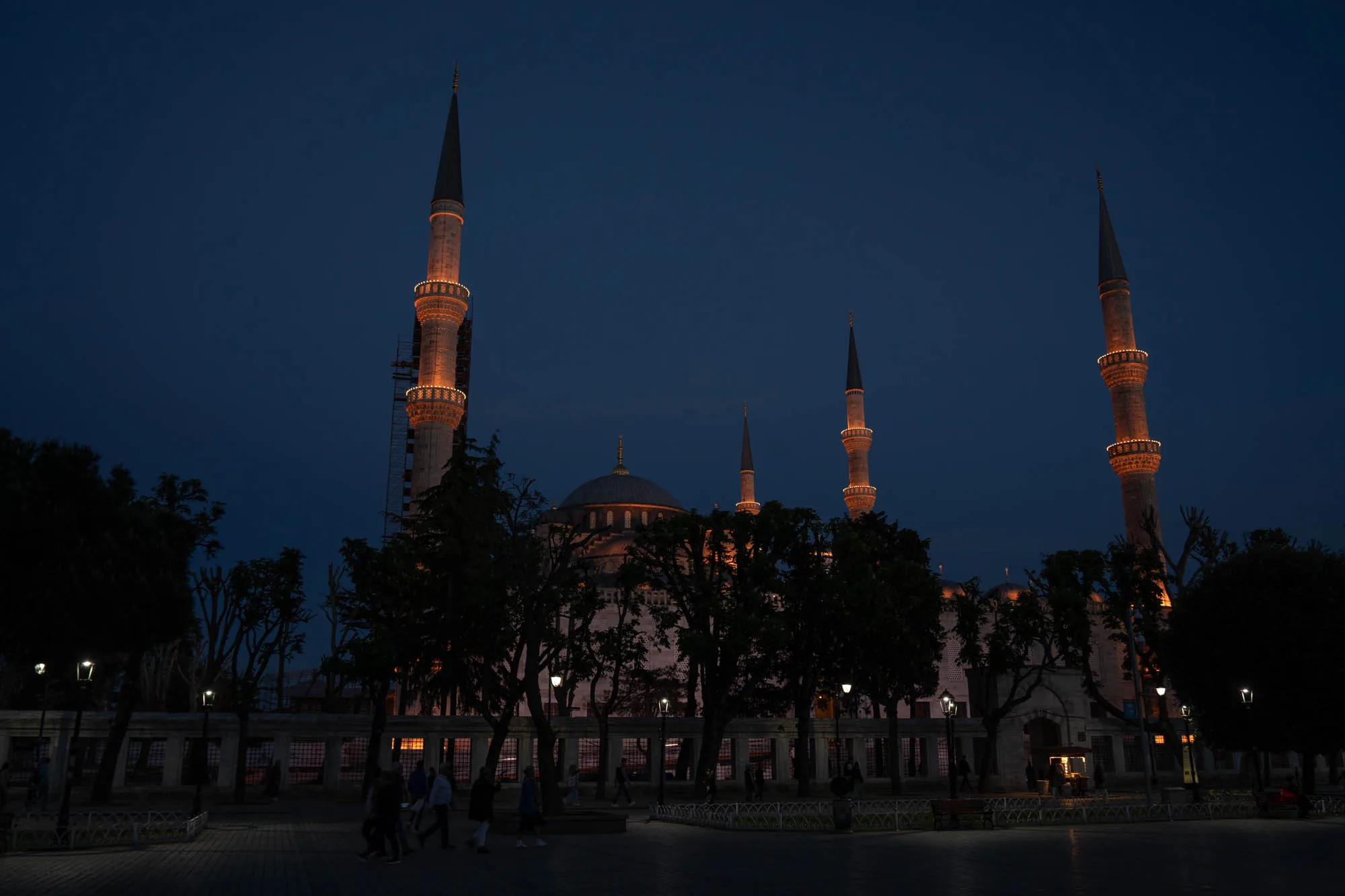 トルコのイスタンブールで撮影された、夜のブルーモスクの写真です。空は深い青色に染まり、モスクはライトアップされています。モスクの尖塔が空にそびえ立ち、オレンジ色の光を放っています。モスクの本体は、シルエットとして浮かび上がり、ドームが特徴的です。前景には、街路樹が並び、人々の姿が小さく見えます。舗装された道には光が反射し、幻想的な雰囲気を醸し出しています。全体的に静かで落ち着いた印象を受ける写真です。