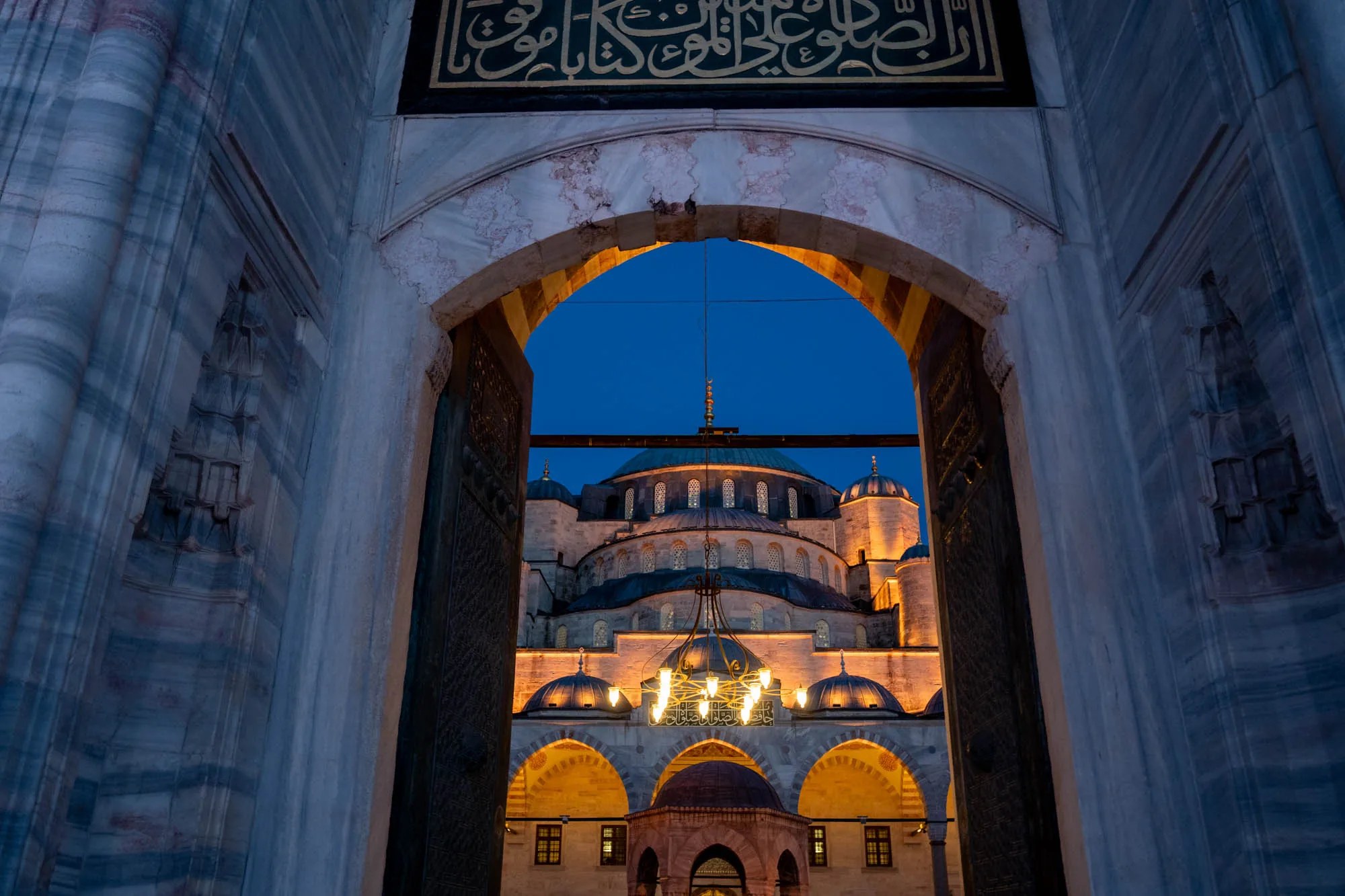 イスタンブールのモスクを、石造りのアーチ型の入り口から捉えた写真です。入り口の石材は、淡い灰色と青色のマーブル模様が美しく、上部にはアラビア文字のような装飾が施されています。アーチの向こうには、ライトアップされたモスクが見え、複数のドームが重なり合っているのが印象的です。モスクの照明は暖色系で、夜空の深い青色とのコントラストが際立っています。アーチ内部には、シャンデリアのような照明が見え、モスク全体の荘厳な雰囲気を高めています。全体的に、静かで神聖な雰囲気が漂う写真です。