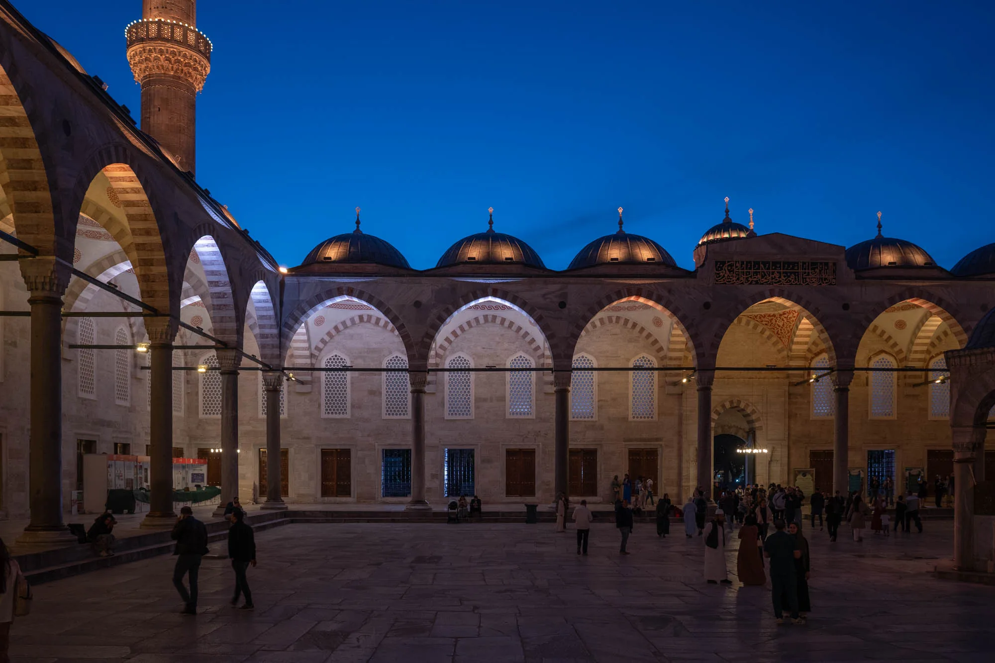 写真はトルコのイスタンブールで撮影された、夜のモスクの様子を捉えています。青みがかった空を背景に、モスクの建物がライトアップされ、美しいコントラストを生み出しています。建物は石造りで、アーチ状の構造が特徴的です。柱が整然と並び、ドーム状の屋根が連なっています。ドームの上にはそれぞれ尖塔のようなものが立っています。建物の壁面には幾何学的な模様や装飾が施されており、歴史と文化を感じさせます。中庭には人々が歩いており、静かで穏やかな雰囲気が漂っています。ライトアップされた建物と、夜空の深い青色が、神秘的で荘厳な雰囲気を醸し出しています。