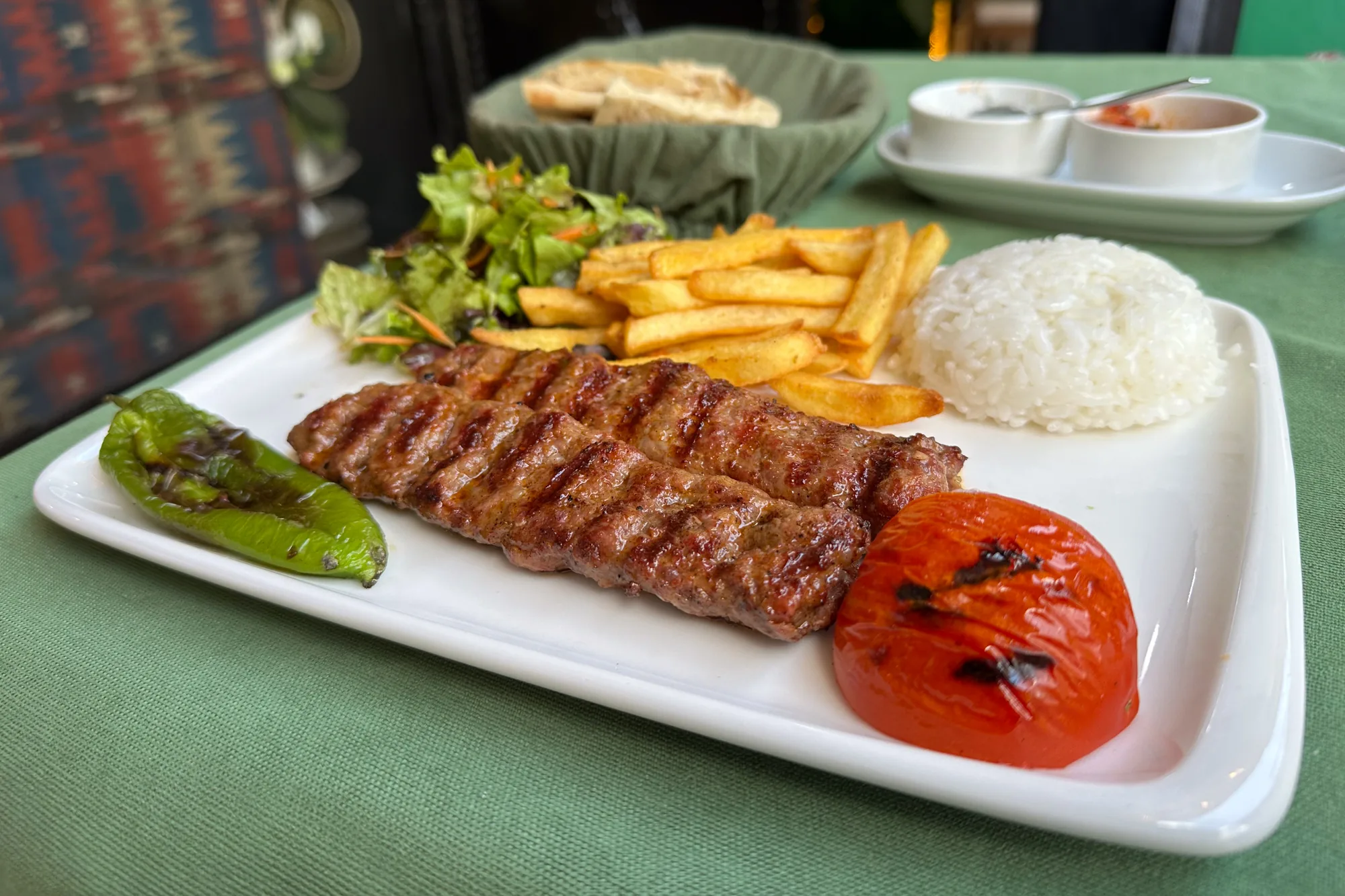写真には、トルコのイスタンブールで撮影されたと思われる料理のクローズアップが写っています。メインはアダナケバブと思われる、串に刺さった挽肉を焼いたもので、焦げ目がついており食欲をそそります。ケバブの横には、焼いた緑色のピーマン、焼いたトマト半分、フライドポテト、ご飯が添えられています。また、サラダも添えられています。白い長方形の皿に盛り付けられており、緑色のテーブルクロスの上に置かれています。背景にはパンが入った籠、ソースが入った小皿が見えます。全体的に、典型的なトルコ料理のセットメニューを写した写真で、レストランの落ち着いた雰囲気と料理のおいしさが伝わってきます。