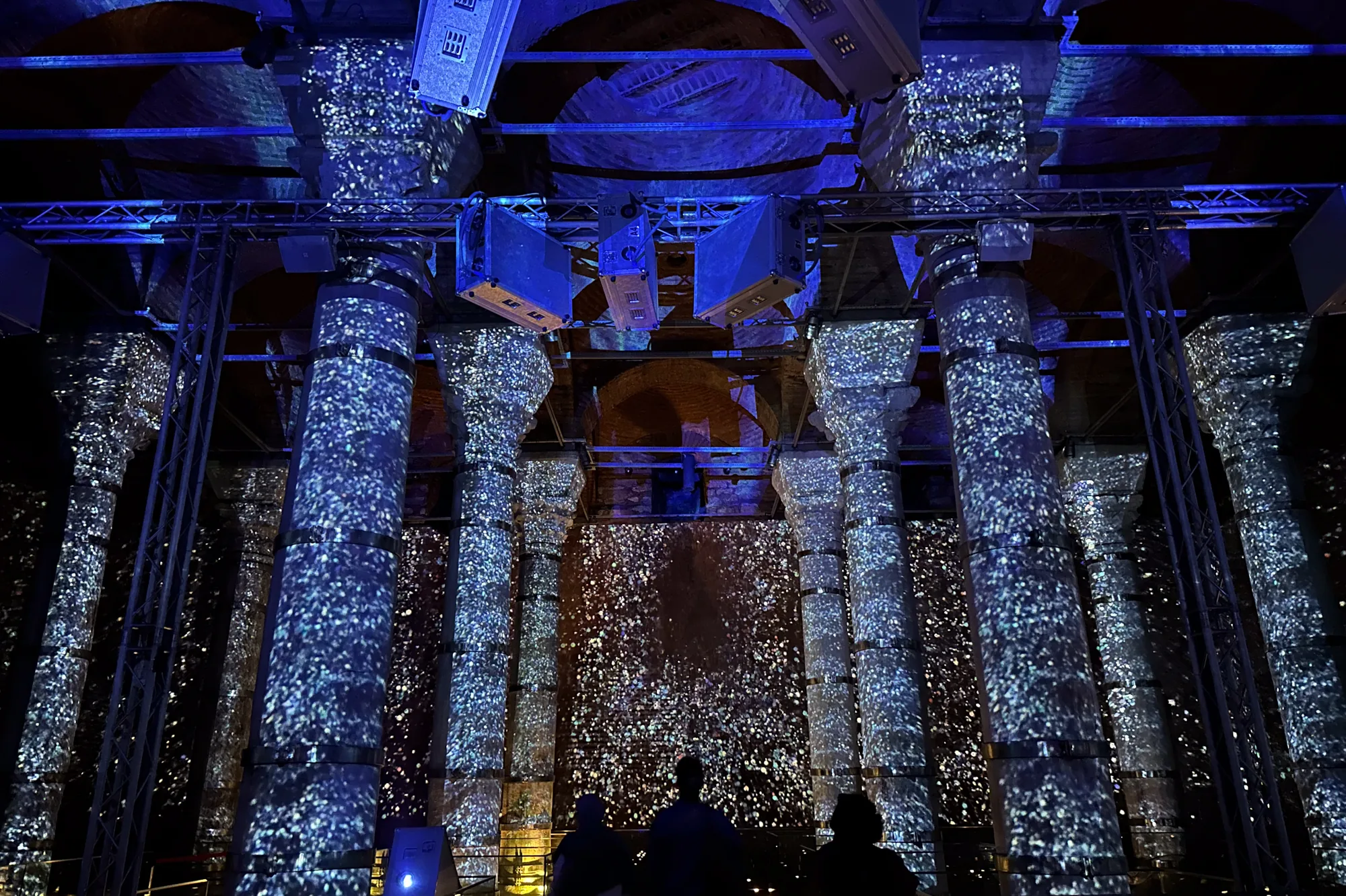 これはトルコのイスタンブールで撮影された写真です。画像には、イスタンブールの地下宮殿の内部が写っています。古代の柱が並び、それらは青みがかった光で照らされています。柱には、まるで星空のような模様が投影されています。天井には、照明機材や構造物が設置されているのが見えます。画像の下部には、鑑賞している人々のシルエットが写っており、空間の広大さを強調しています。全体的に、神秘的で幻想的な雰囲気を醸し出しています。