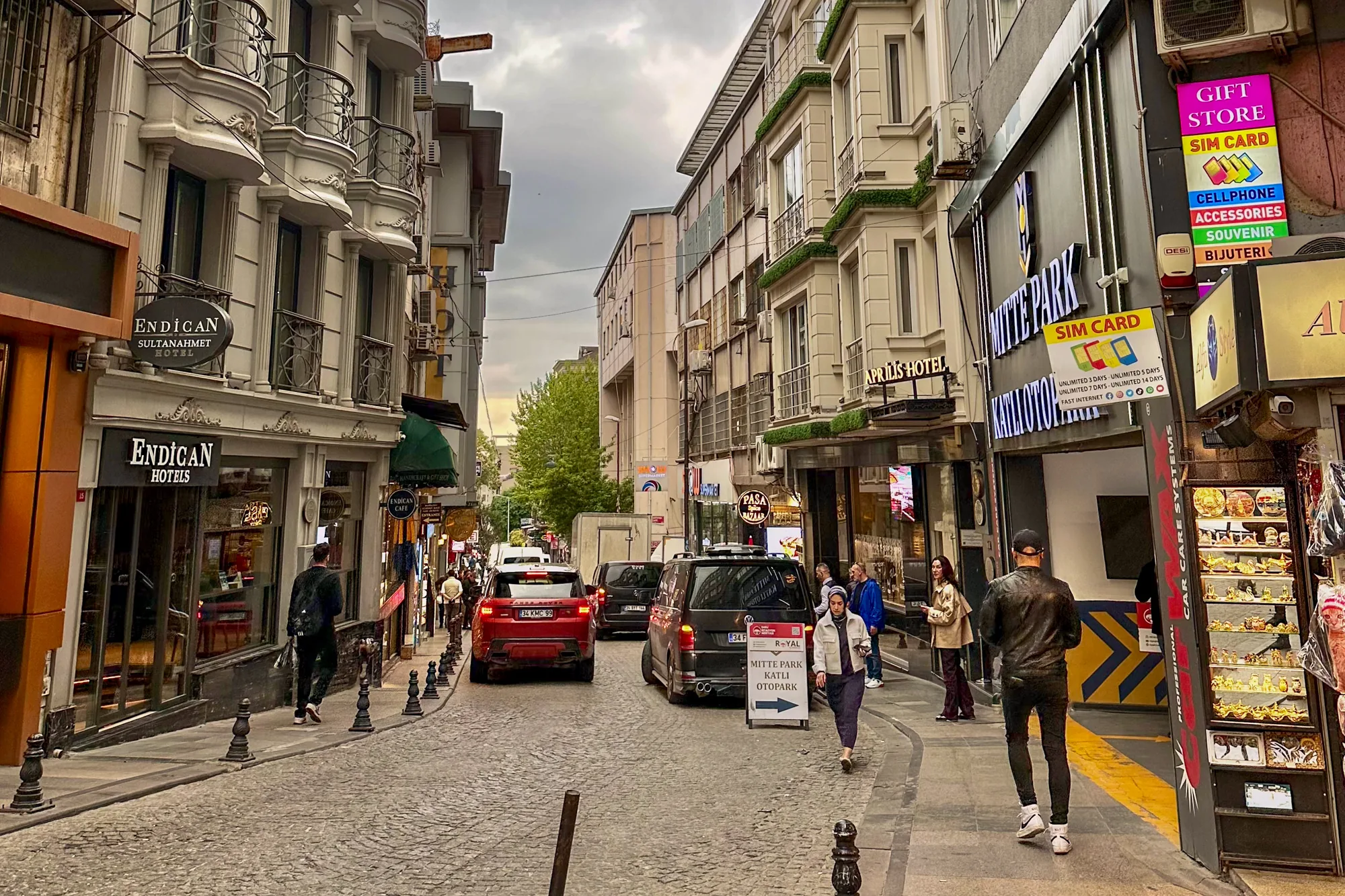 写真はトルコのイスタンブールで撮影されたもので、石畳の道が特徴的な街並みが写っています。通りの両側にはホテルやショップが立ち並び、観光客や地元の人々が行き交っています。左側には「ENDICAN SULTANAHMET HOTEL」と書かれた看板が見え、右側にはSIMカードや携帯アクセサリーなどを販売する店があります。道路には数台の車が駐車しており、行き交う人々の様子が見られます。空は曇り空で、全体的に落ち着いた雰囲気です。写真に写っている建物のデザインはヨーロッパ風で、イスタンブールの歴史的な魅力が感じられます。