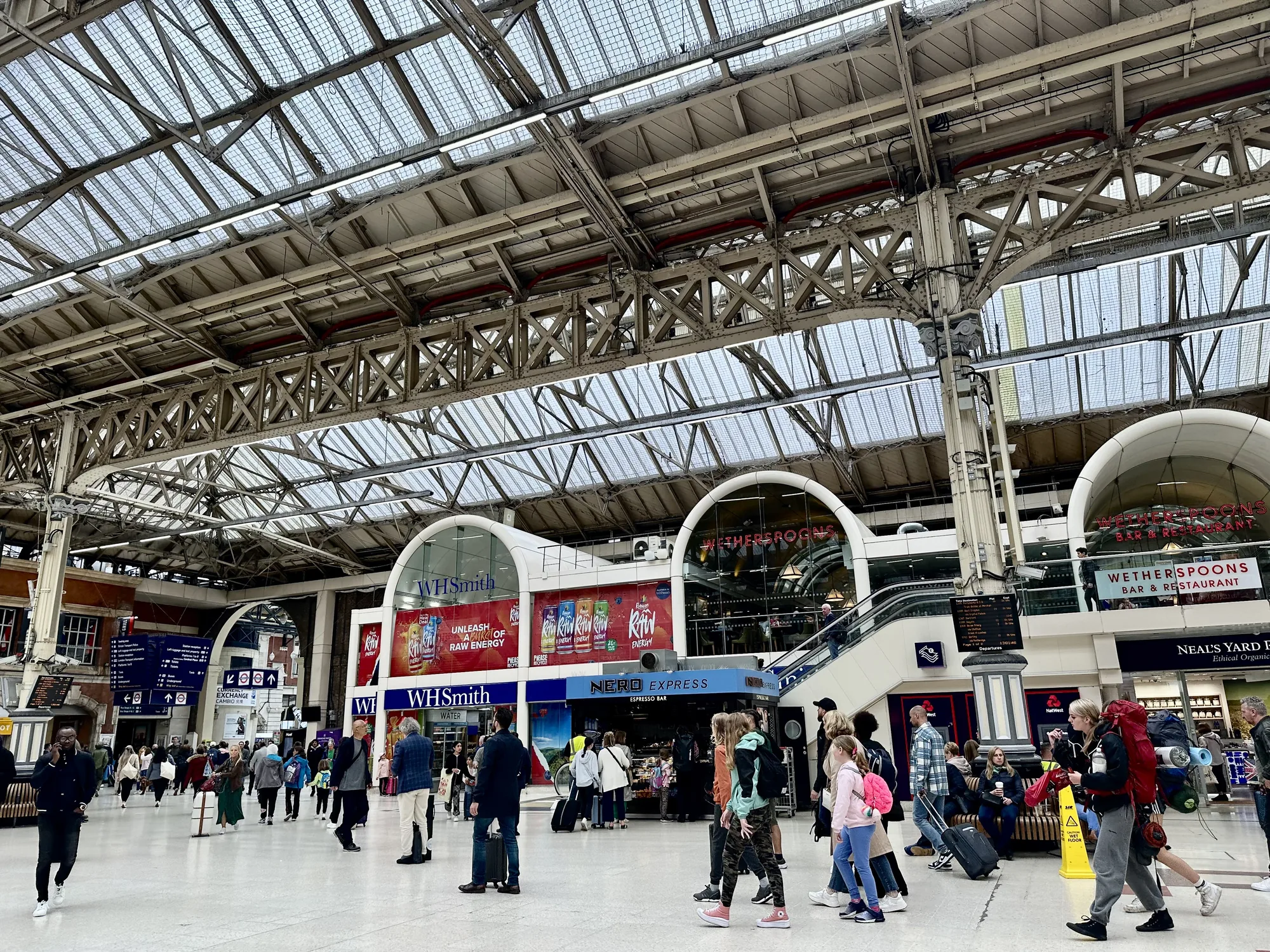 写真は、イギリス・ロンドンの駅構内を捉えたものです。高い天井はガラス張りで、複雑な鉄骨構造が印象的です。多くの人々が行き交っており、旅行者や通勤客などが混ざり合っている様子が伺えます。構内には、WHSmith、Nero Express、Wetherspoonsなどの店舗が並んでおり、活気があります。床は磨かれており、光を反射して明るい印象を与えています。全体的に、歴史を感じさせる建築と現代的な店舗が共存する、ロンドンの駅の賑やかな雰囲気が伝わってくる写真です。