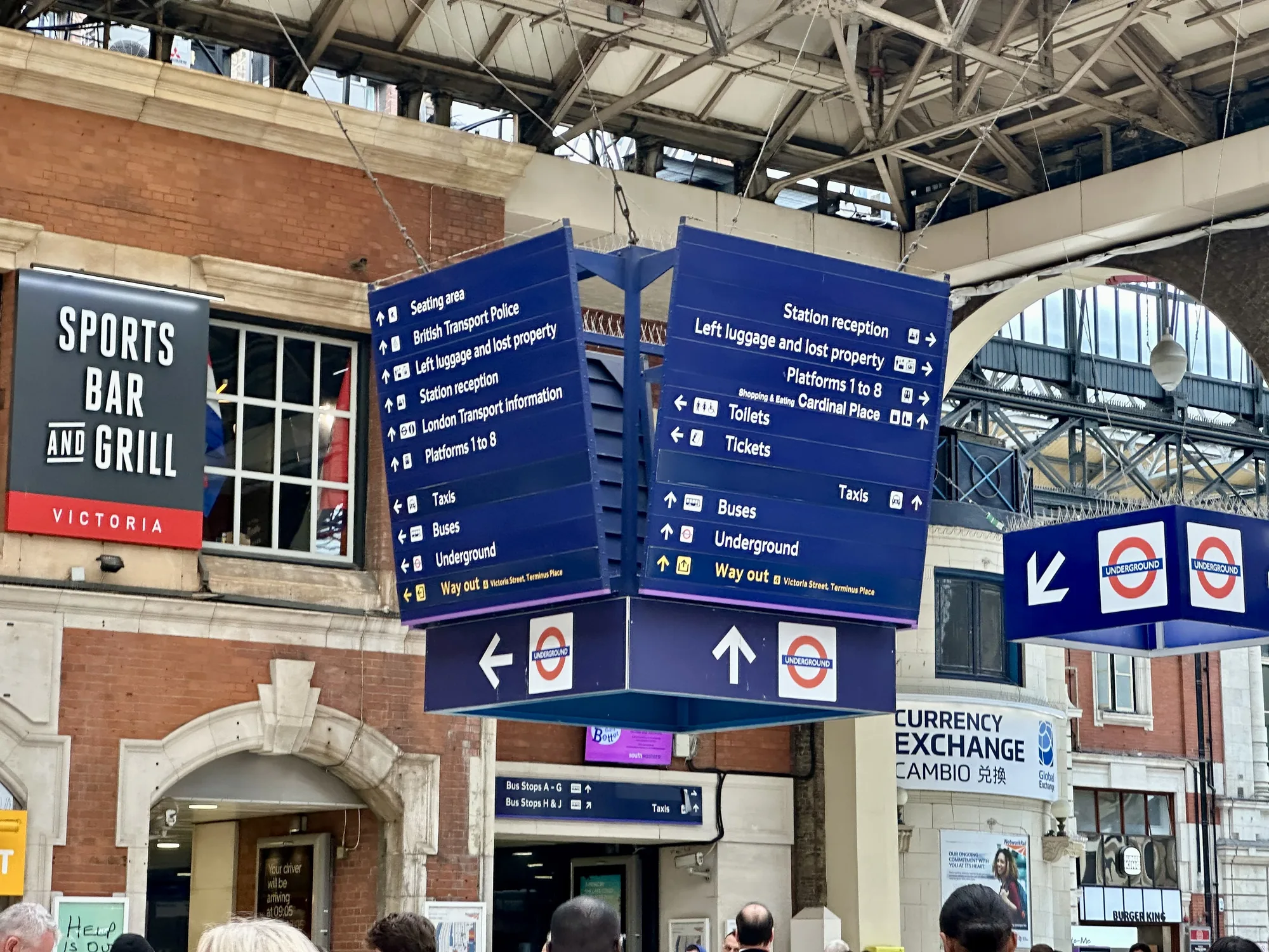 写真は、イギリスのロンドンの駅構内で撮影されたものです。中央には、天井から吊り下げられた大きな青色の案内標識が写っています。標識には、駅の設備や目的地への方向を示す矢印や文字が書かれており、具体的には「座席エリア」、「英国運輸警察」、「手荷物預かり所」、「駅受付」、「ロンドン交通情報」、「1番から8番のプラットフォーム」、「タクシー」、「バス」、「地下鉄」、「出口」などが確認できます。標識の下部には、ロンドンの地下鉄を示す赤い円形のロゴマークが描かれています。  背景には、レンガ造りの建物が見え、一部には「SPORTS BAR AND GRILL VICTORIA」と書かれた看板や、両替所の看板、バーガーキングの看板なども確認できます。これらの看板は、駅周辺の商業施設を示しています。また、写っている人々は駅を利用する人々であると考えられます。天井部分は複雑な鉄骨構造で、歴史を感じさせる雰囲気を醸し出しています。全体的に、ロンドンの主要な駅構内のにぎやかさと機能性を伝える写真です。