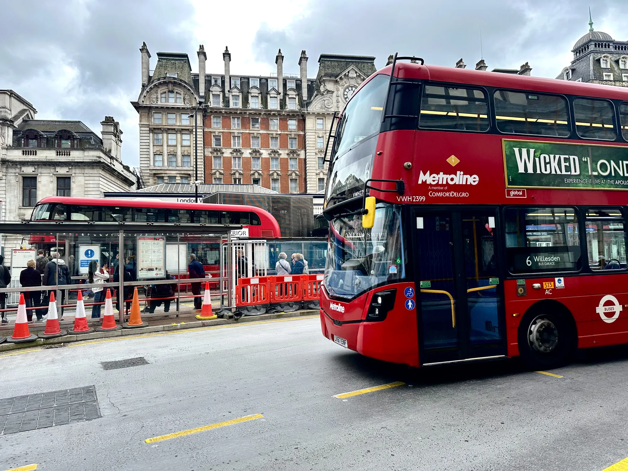 写真はイギリスのロンドンの街並みを捉えています。手前には赤い二階建てバスが大きく写っており、"Metroline"というバス会社のロゴや、ミュージカル「Wicked」の広告が見えます。バスの行き先番号は6番で、Willesdenのバスガレージ方面行きです。バスのそばには歩行者用の黄色い点線が引かれた道路があります。  バスの後ろにはバス停があり、数人の人々がバスを待っている様子がうかがえます。バス停の手前には赤と白の三角コーンが並べられています。  背景には、歴史を感じさせる趣のある建物が写っており、時計台や装飾的な屋根が見えます。空は曇っており、全体的に落ち着いた雰囲気の写真です。